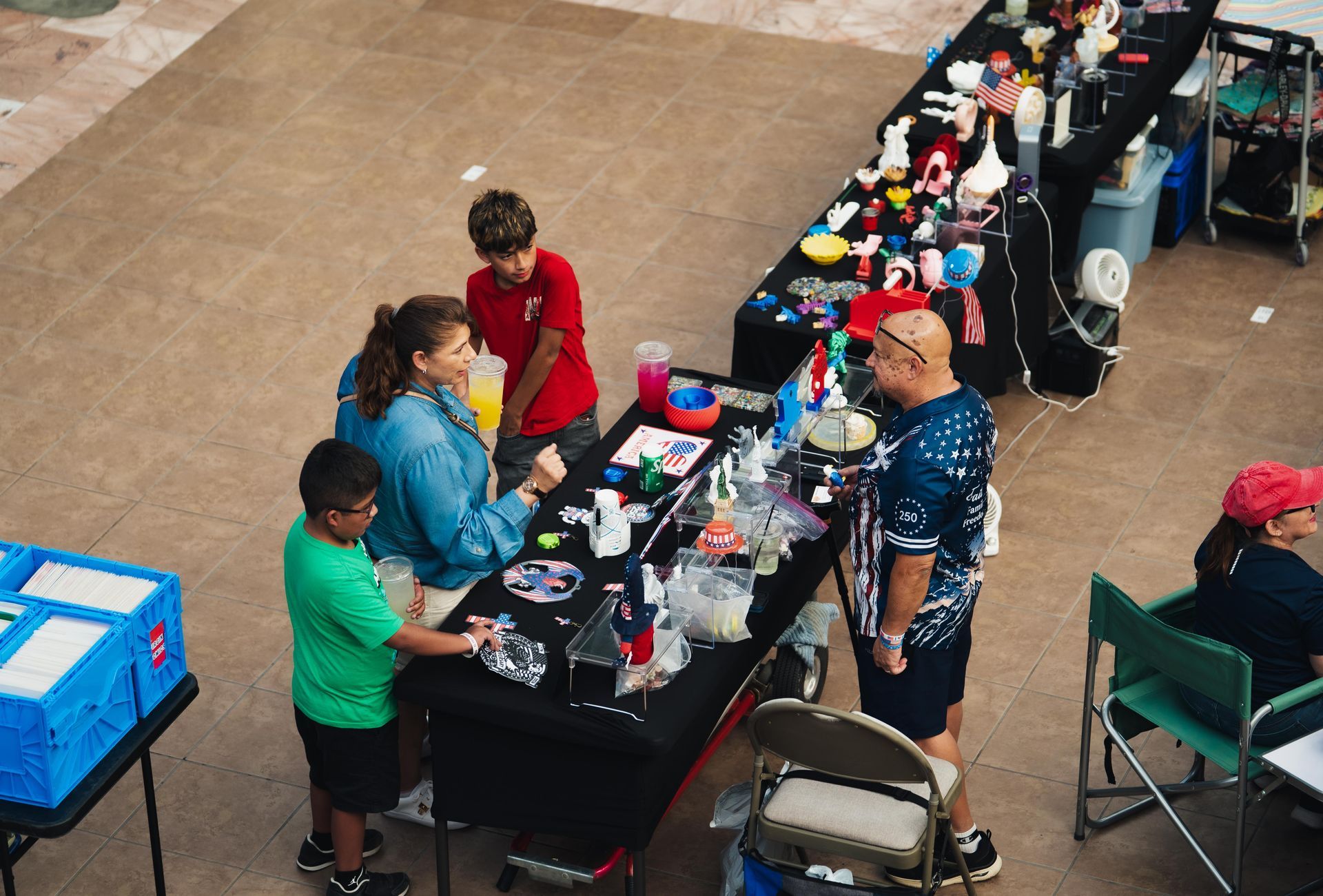 People seated at a long vendor table with colorful items and supplies in an outdoor plaza