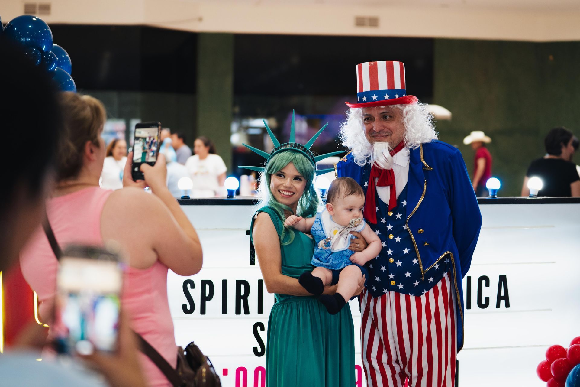 Costumed man and woman holding a child at a festive indoor event with balloons and an American flag outfit