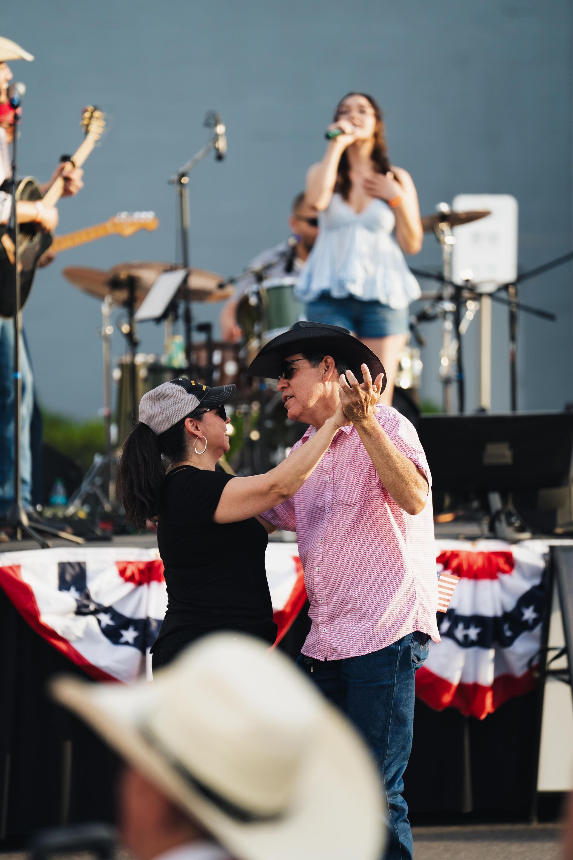 Two dancers in a cowboy hat and pink shirt twirl onstage before a band with patriotic bunting.