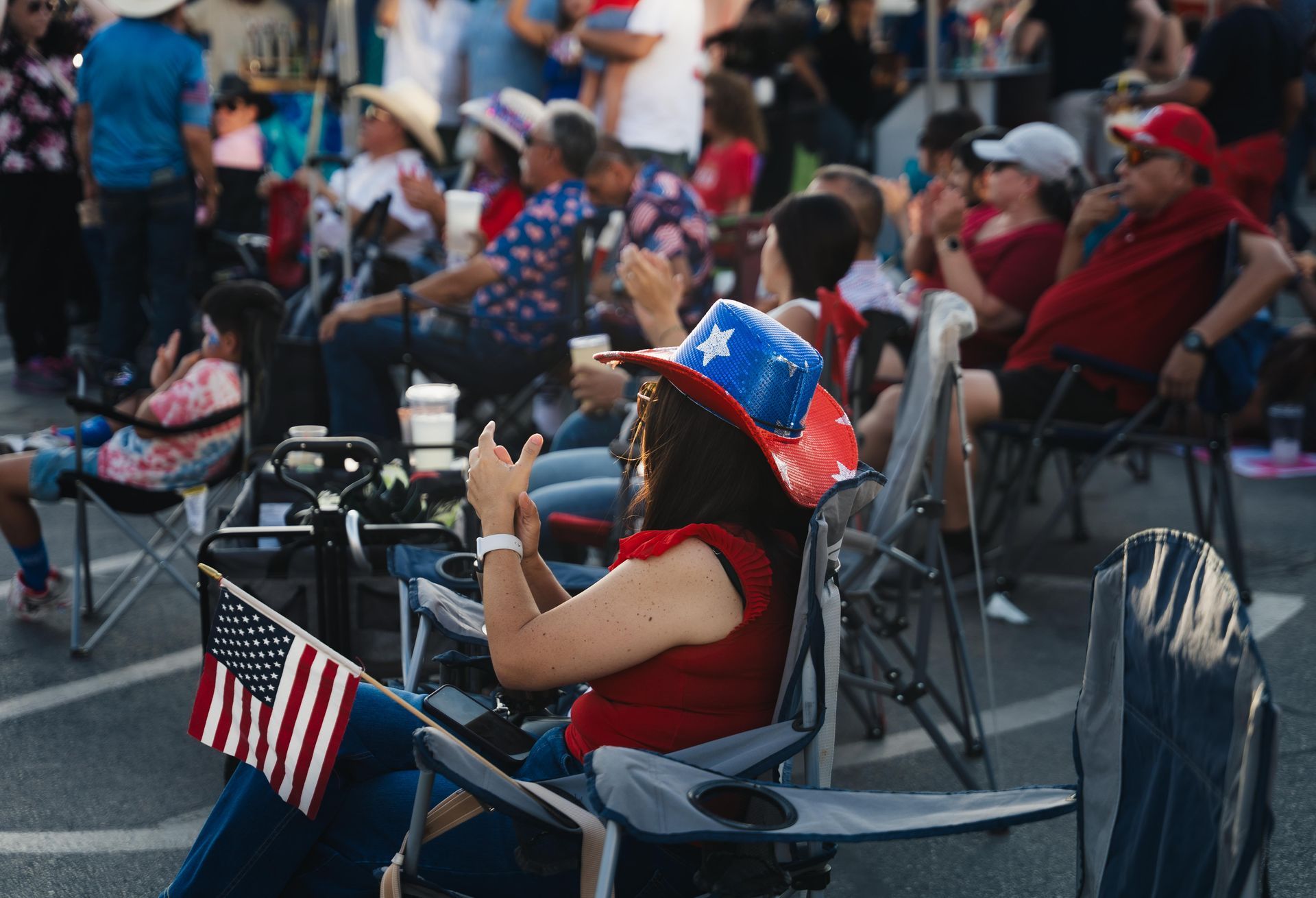 People seated outdoors at a patriotic event, some wearing red, white, and blue hats and holding small American flags.