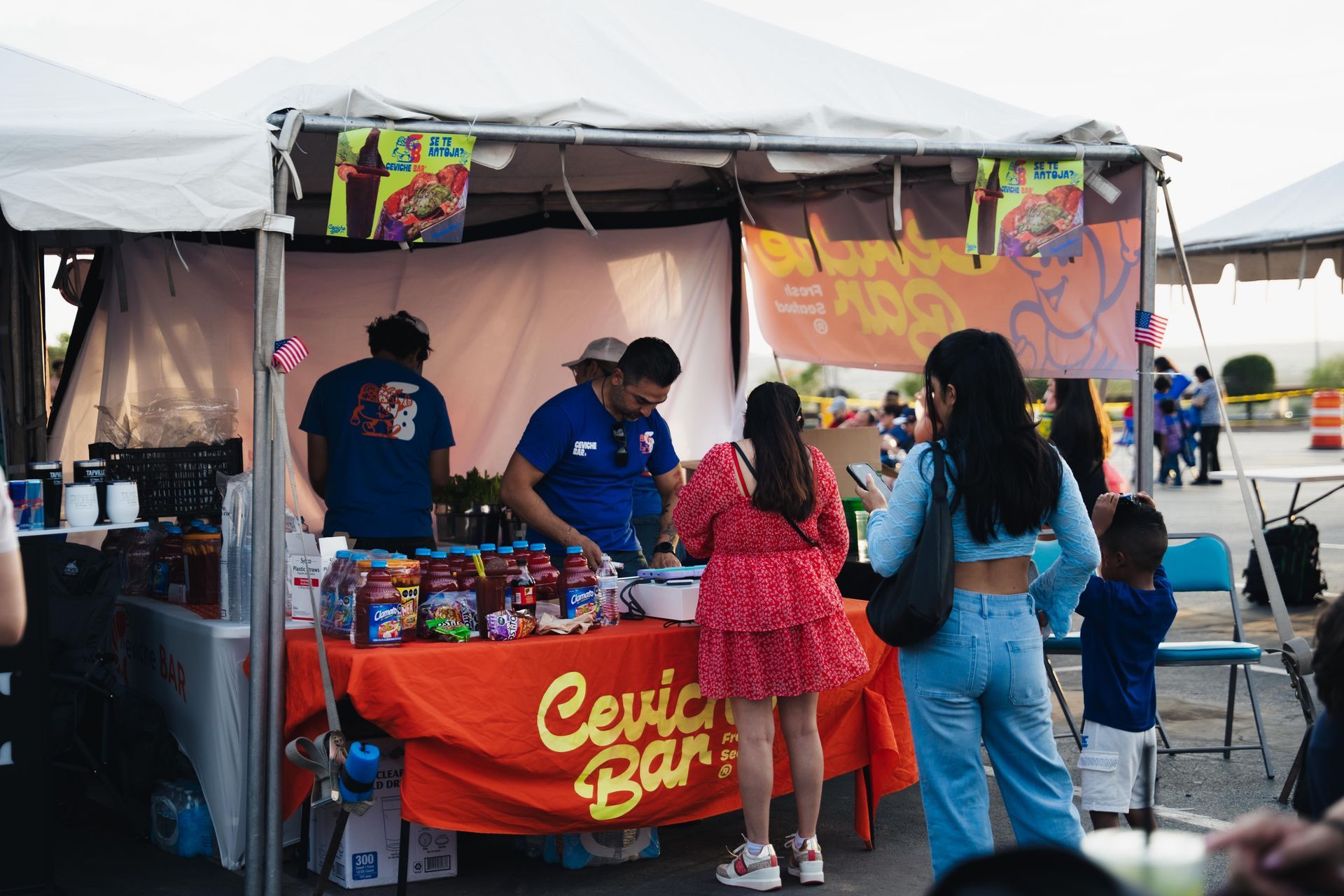 Outdoor food stall under a white canopy, with two vendors serving customers beside a red table banner.