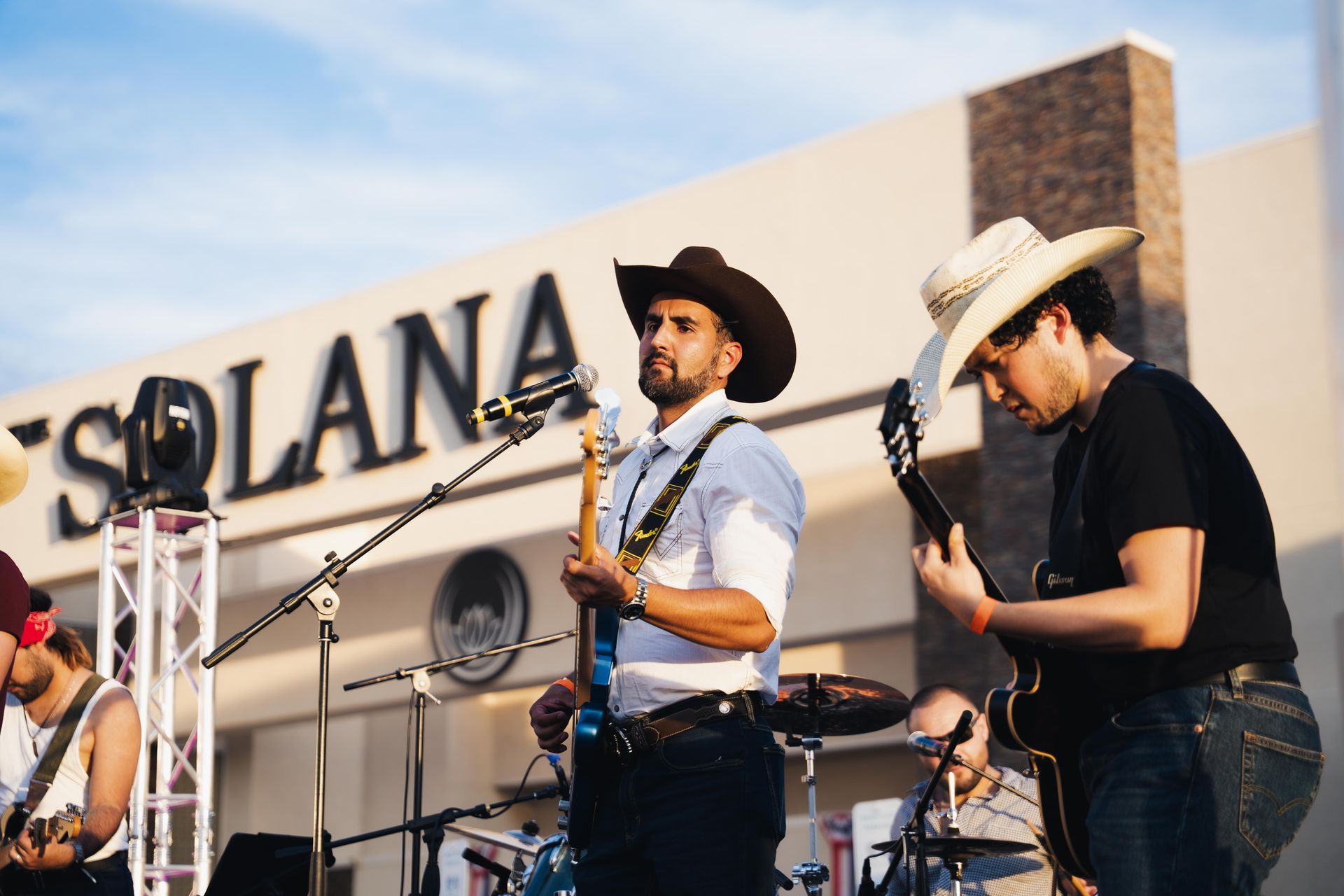 Live band performing outdoors in front of a Solana sign, with musicians playing guitar and hat-wearing singer at microphones.
