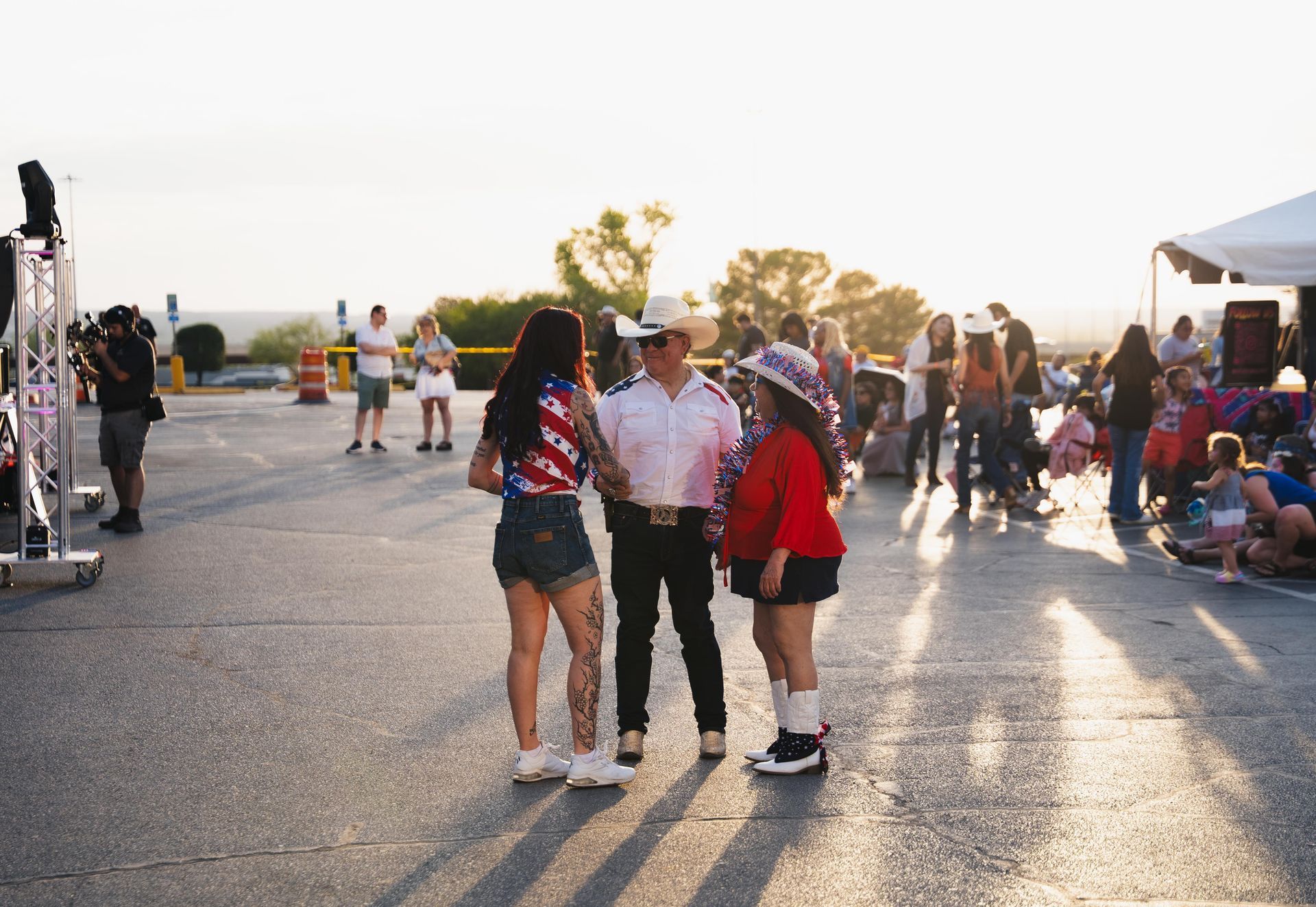 Three people talking in a sunlit parking lot at an outdoor event, with crowds and tents in the background.