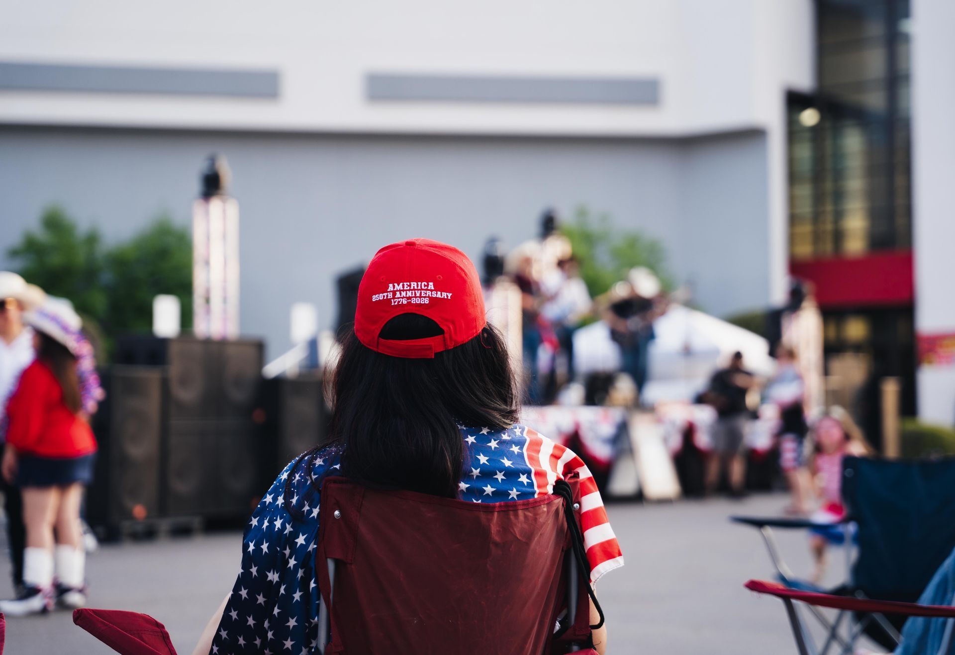 Person in red cap and American flag shirt seated at an outdoor event with a stage in the background