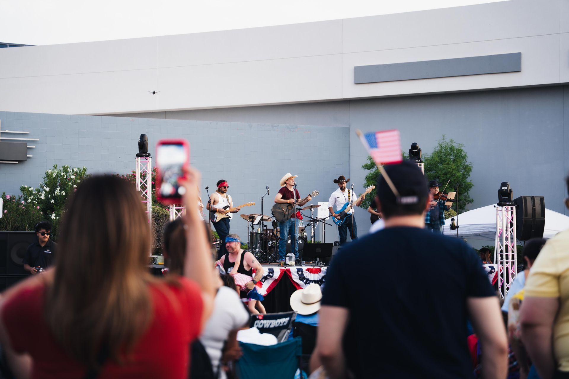 Outdoor concert with a band onstage, seen from a crowd holding up phones in front of a gray building.