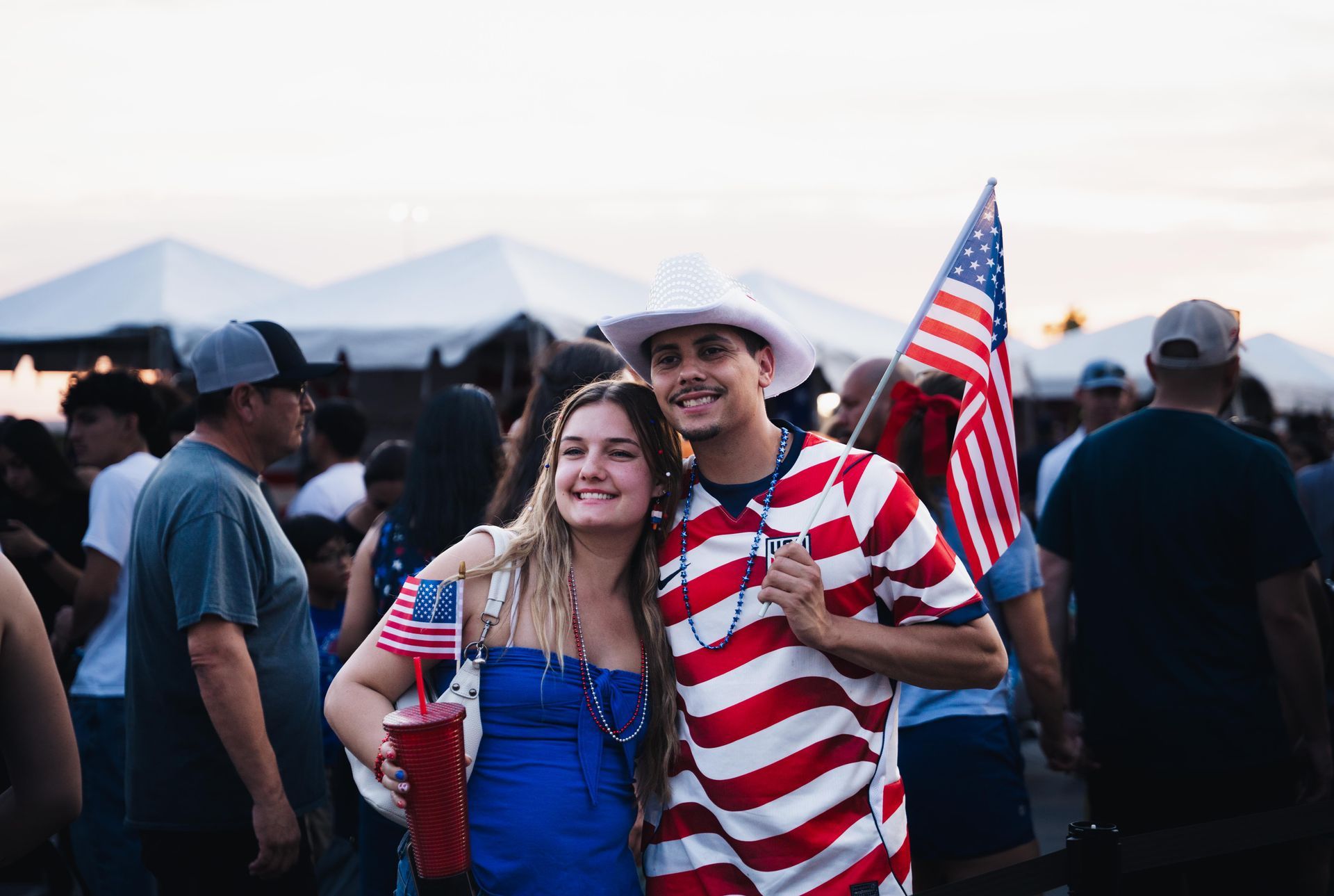 Two people posing at an outdoor event, one in a red-and-white striped outfit holding an American flag.
