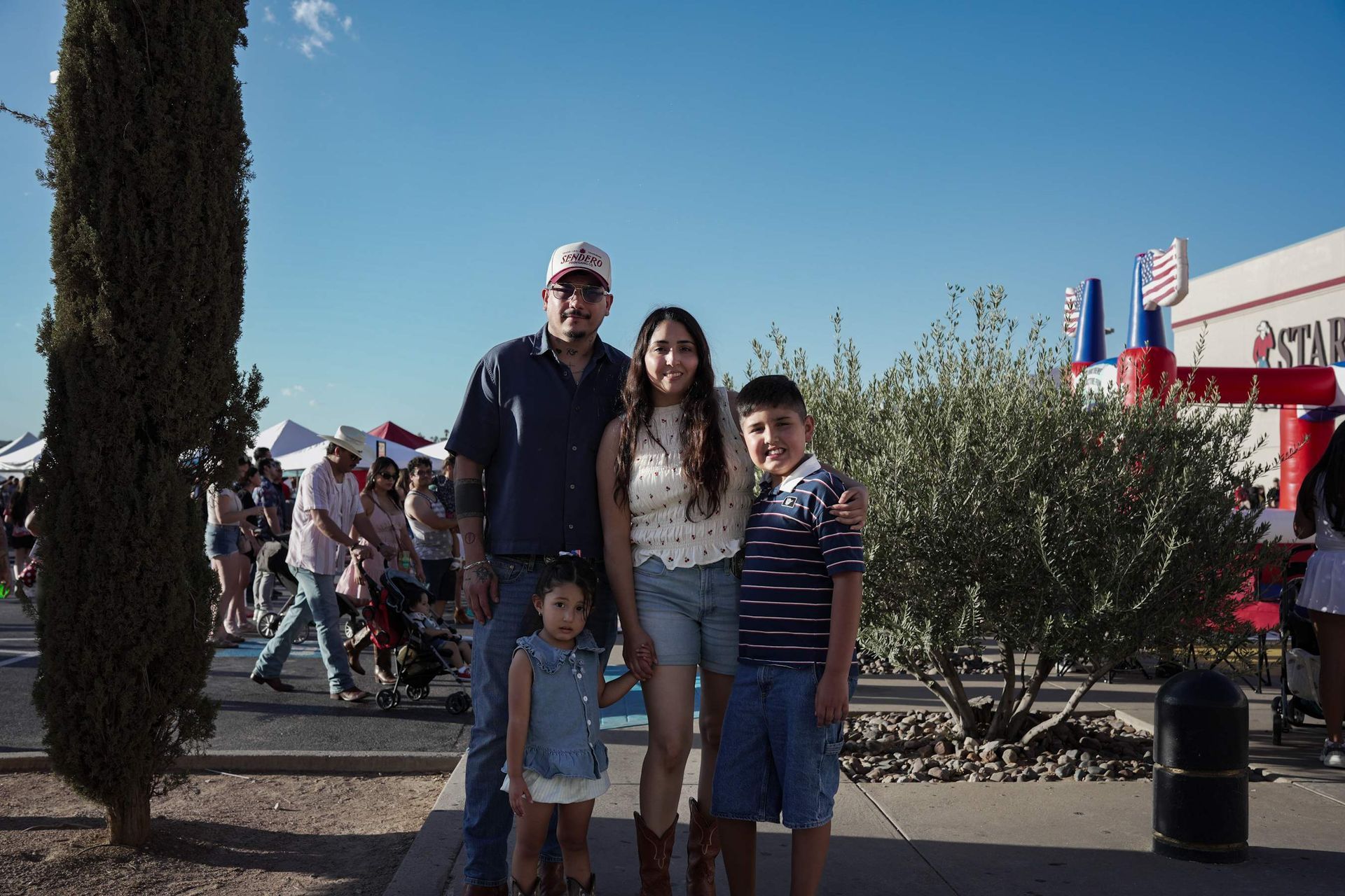 Family posing outdoors at a fair, with a tall tree, blue sky, and colorful tents in the background.