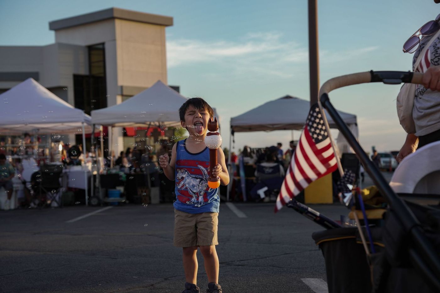 Child smiling at an outdoor evening event, with tents and an American flag in the background.