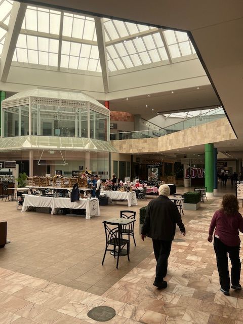 Spacious indoor mall atrium with a skylight, glass elevator, kiosk tables, and shoppers walking through.