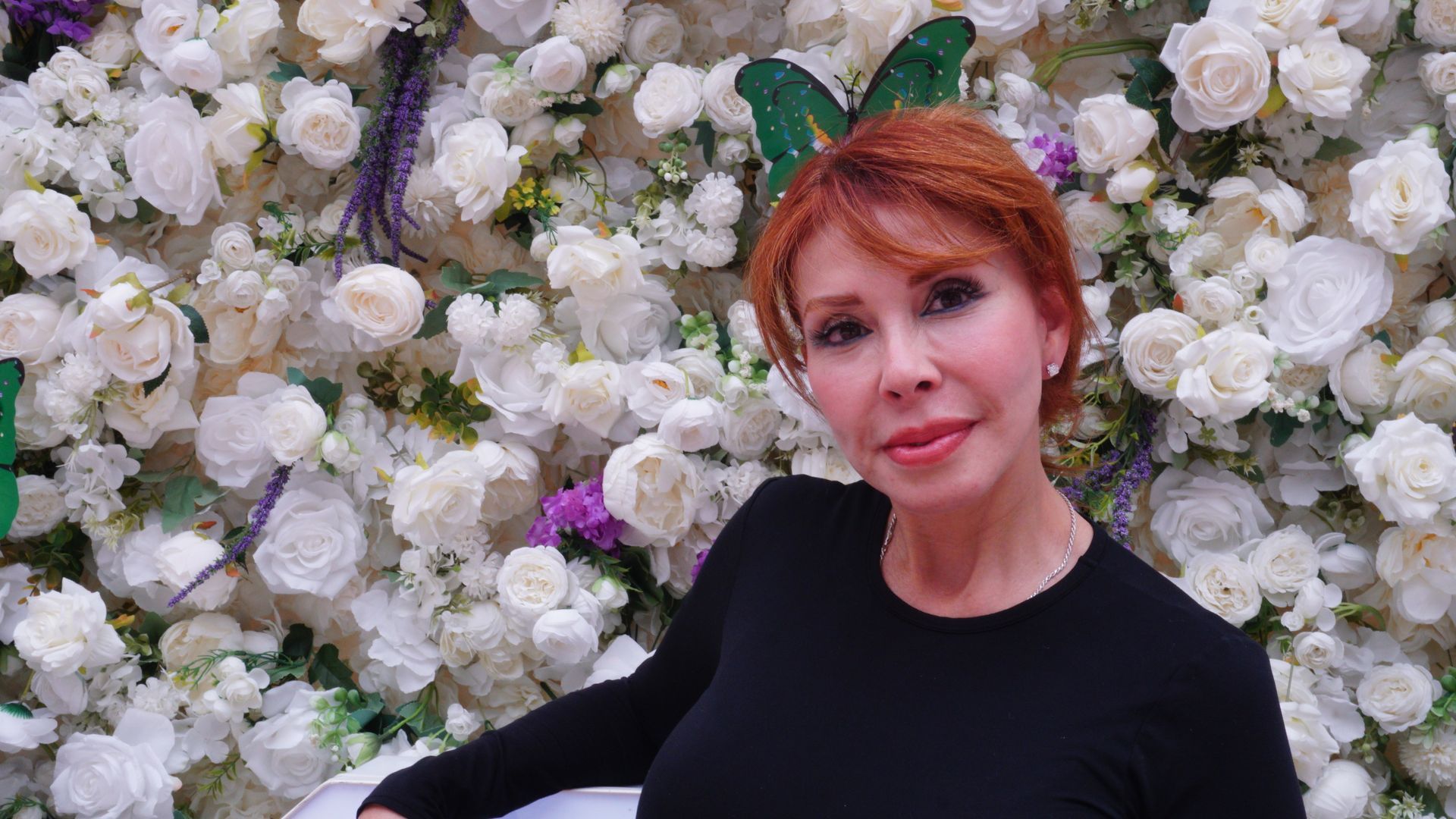 Woman in a black top posing before a wall of white flowers, with a soft smile.