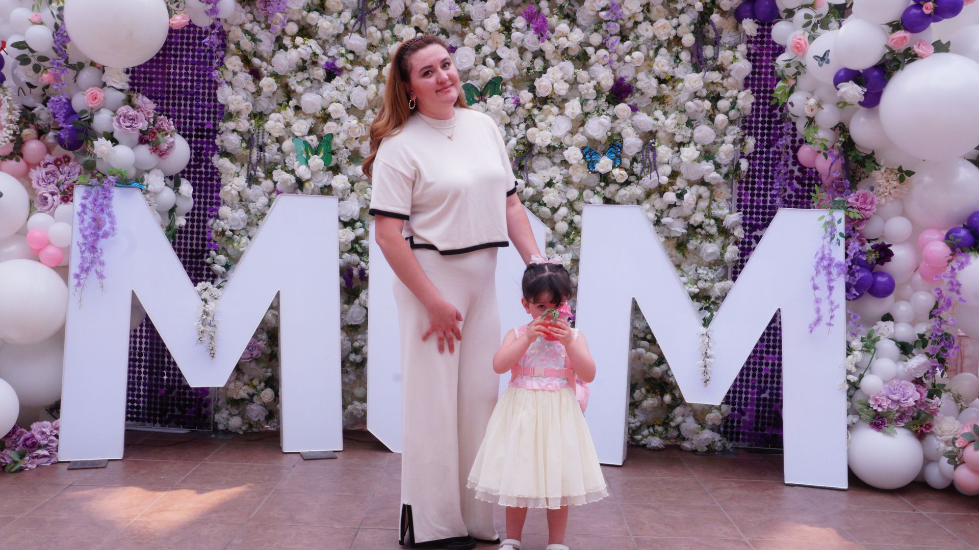 Woman and child posing before a floral “MOM” backdrop with white balloons at a celebration