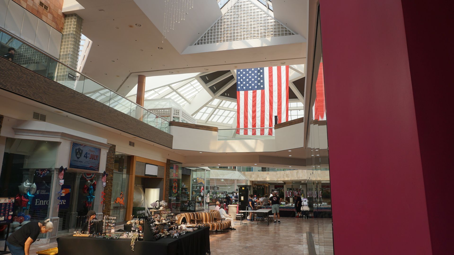 Indoor shopping mall atrium with a large American flag hanging above storefronts and seating areas