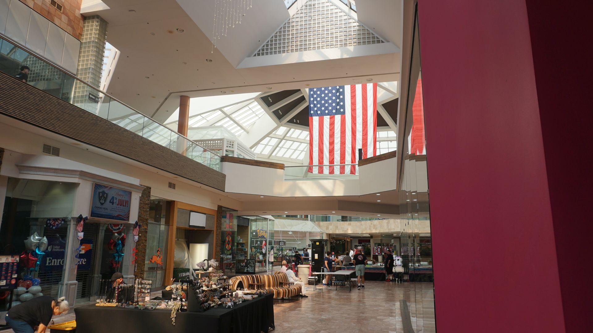 Indoor shopping mall atrium with a large American flag hanging from the ceiling and shoppers below