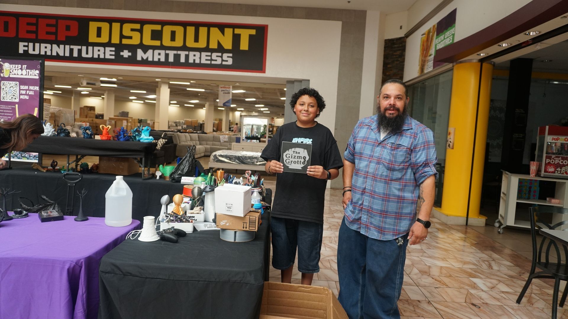 Two men stand outside a Deep Discount Furniture & Mattress storefront beside a table of items.