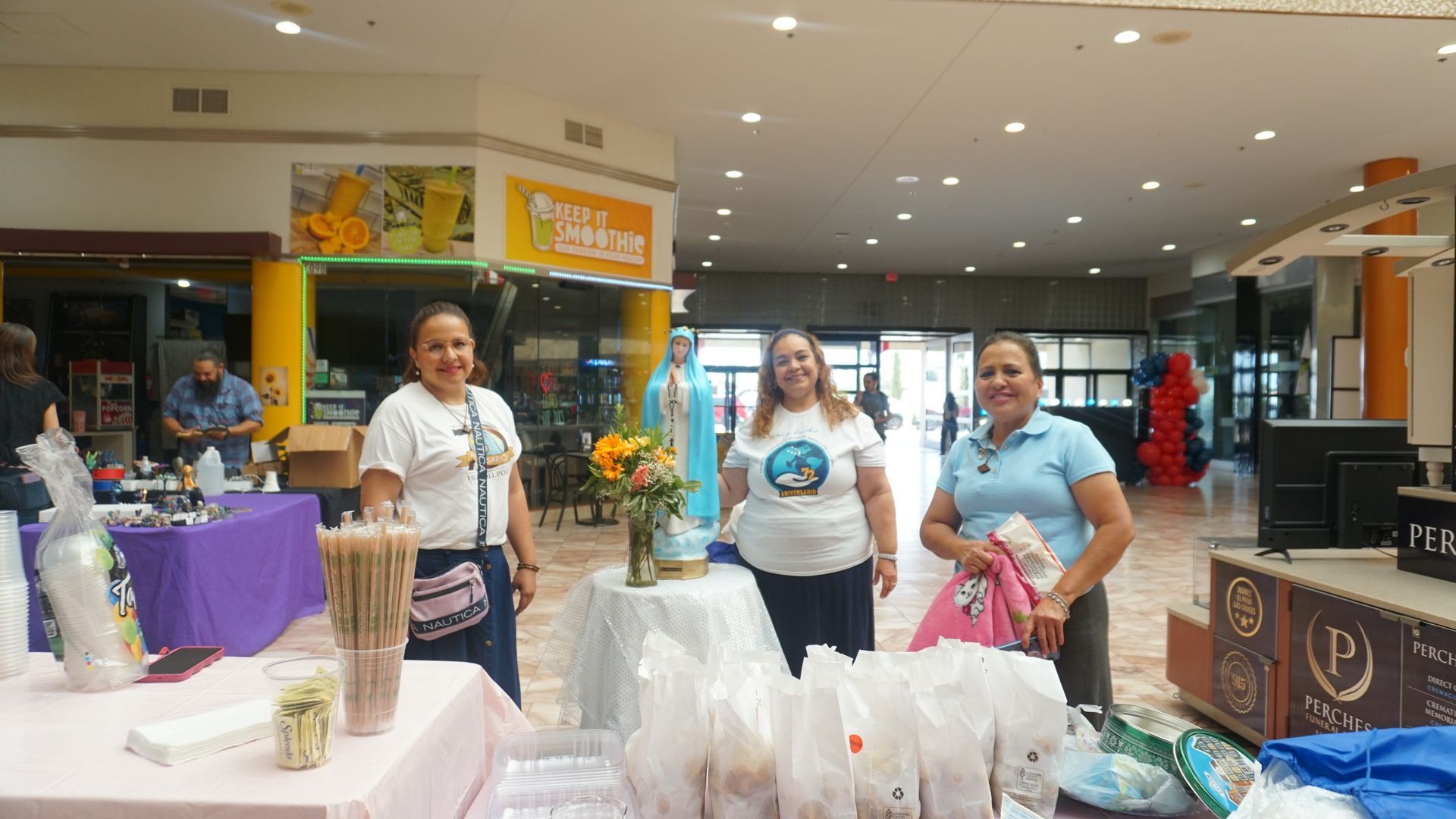 People standing at a mall booth with tables of goods and plastic bags in a bright indoor market.