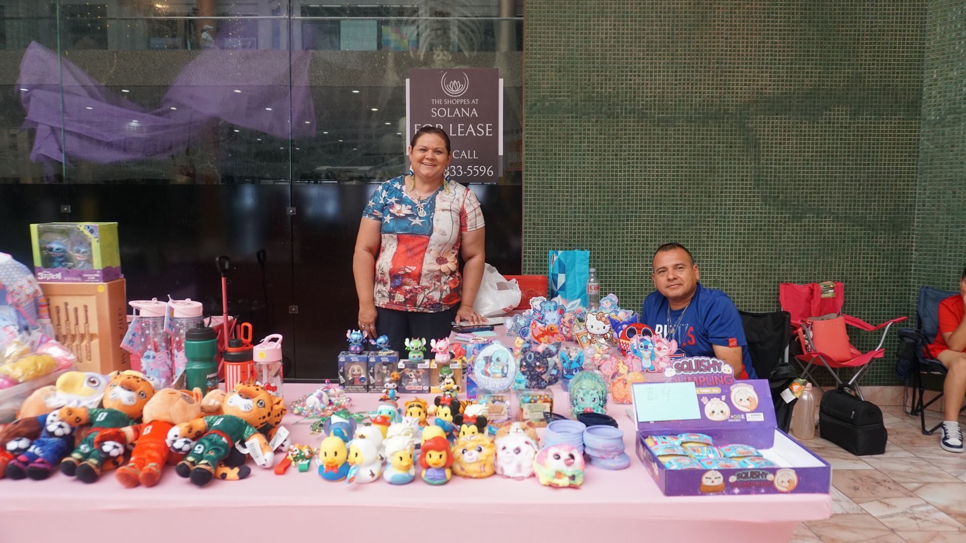 Vendor smiling behind a table of colorful stuffed toys at an outdoor market stall