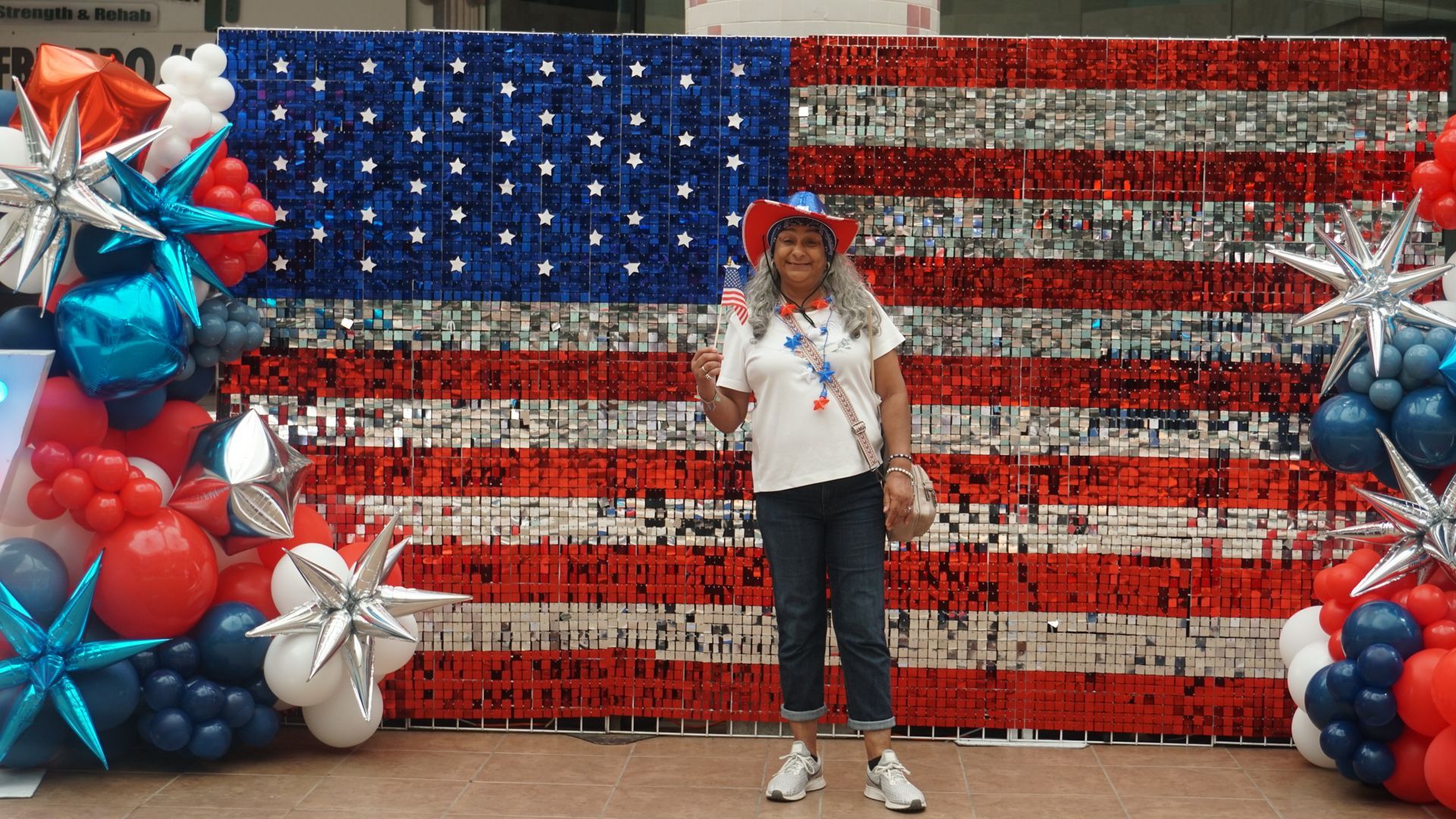 Person posing before a red, white, and blue patriotic backdrop with stars, balloons, and pinwheel decorations