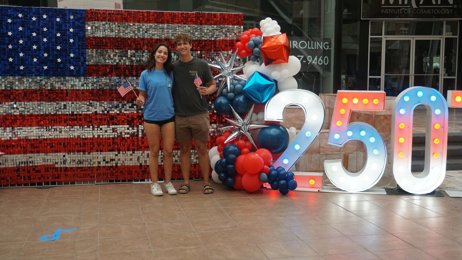 Two people posing in front of a U.S. flag mural with balloons and illuminated “250” numbers.