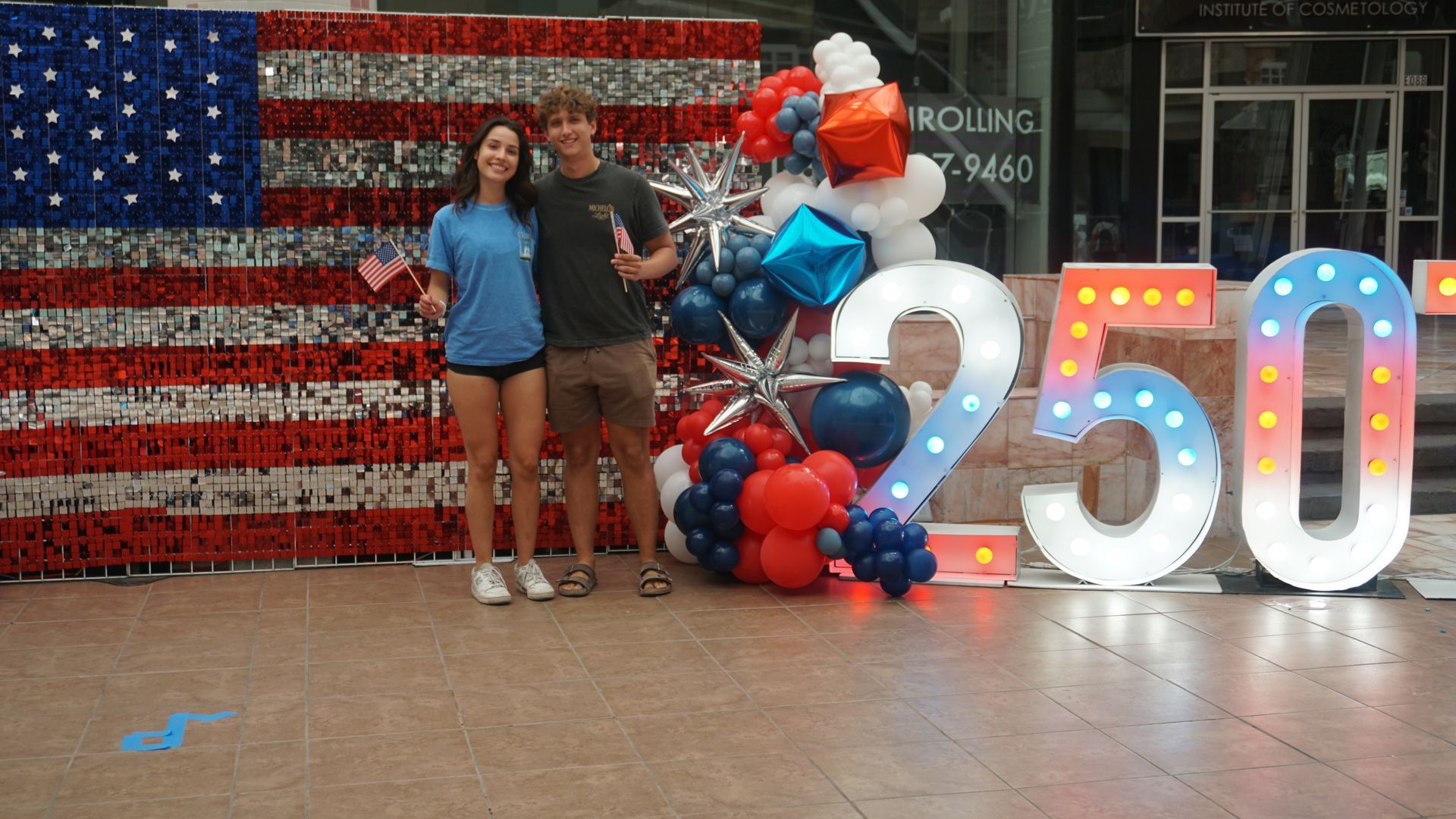 Two people posing beside a red, white, and blue balloon display and lit “250” numbers against an American flag backdrop