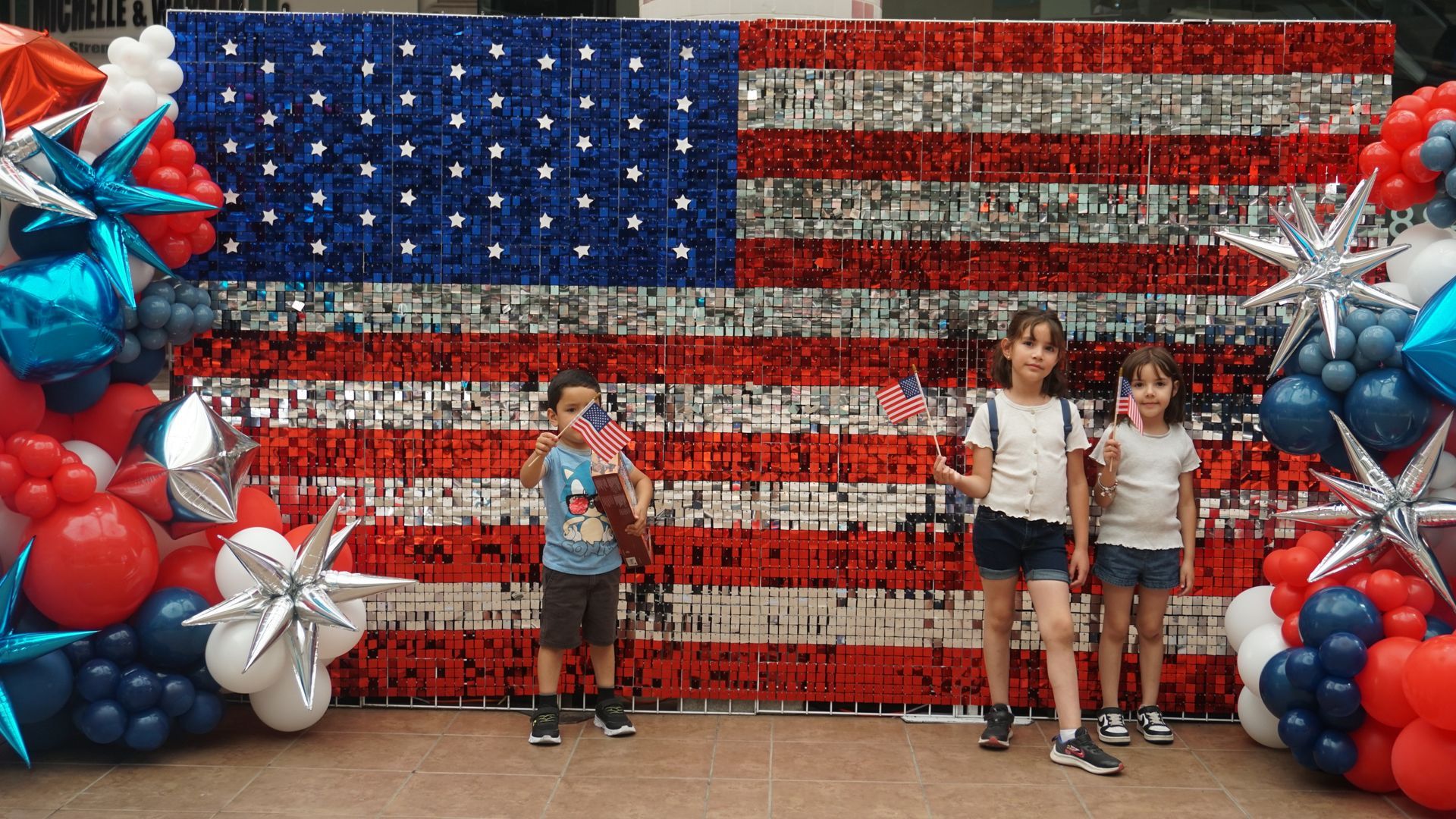 Children posing in front of a large American flag backdrop with red, white, and blue balloons