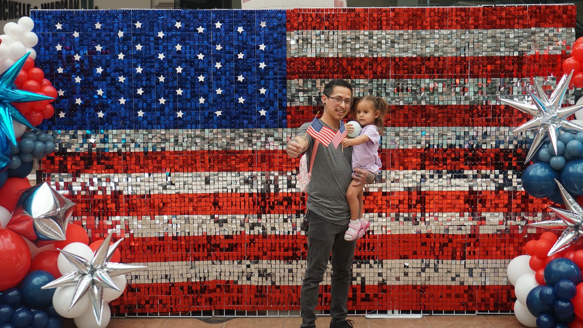 Person holding a child before a large American flag made of flowers, with red, white, and blue balloon decorations.