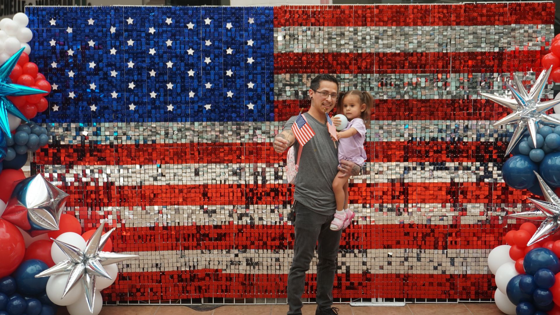 Person holding a child in front of a large American flag display with red, white, and blue balloon decorations