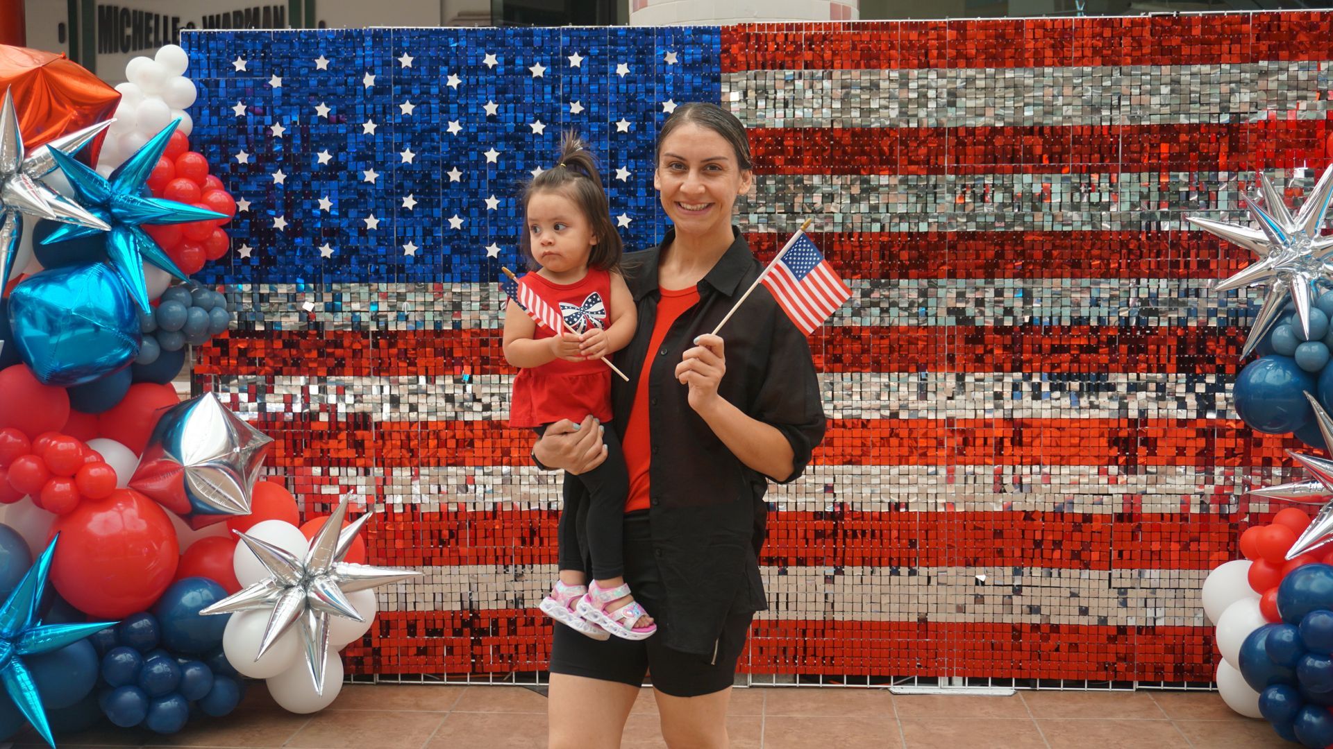 Woman holding a child in front of a red, white, and blue American flag balloon backdrop with star decorations