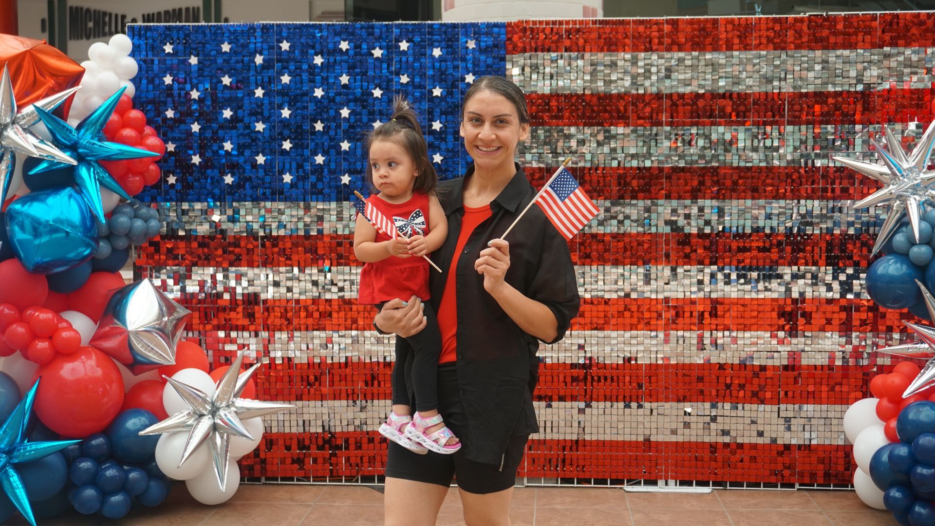 Woman holding a child in front of a red, white, and blue American flag balloon backdrop
