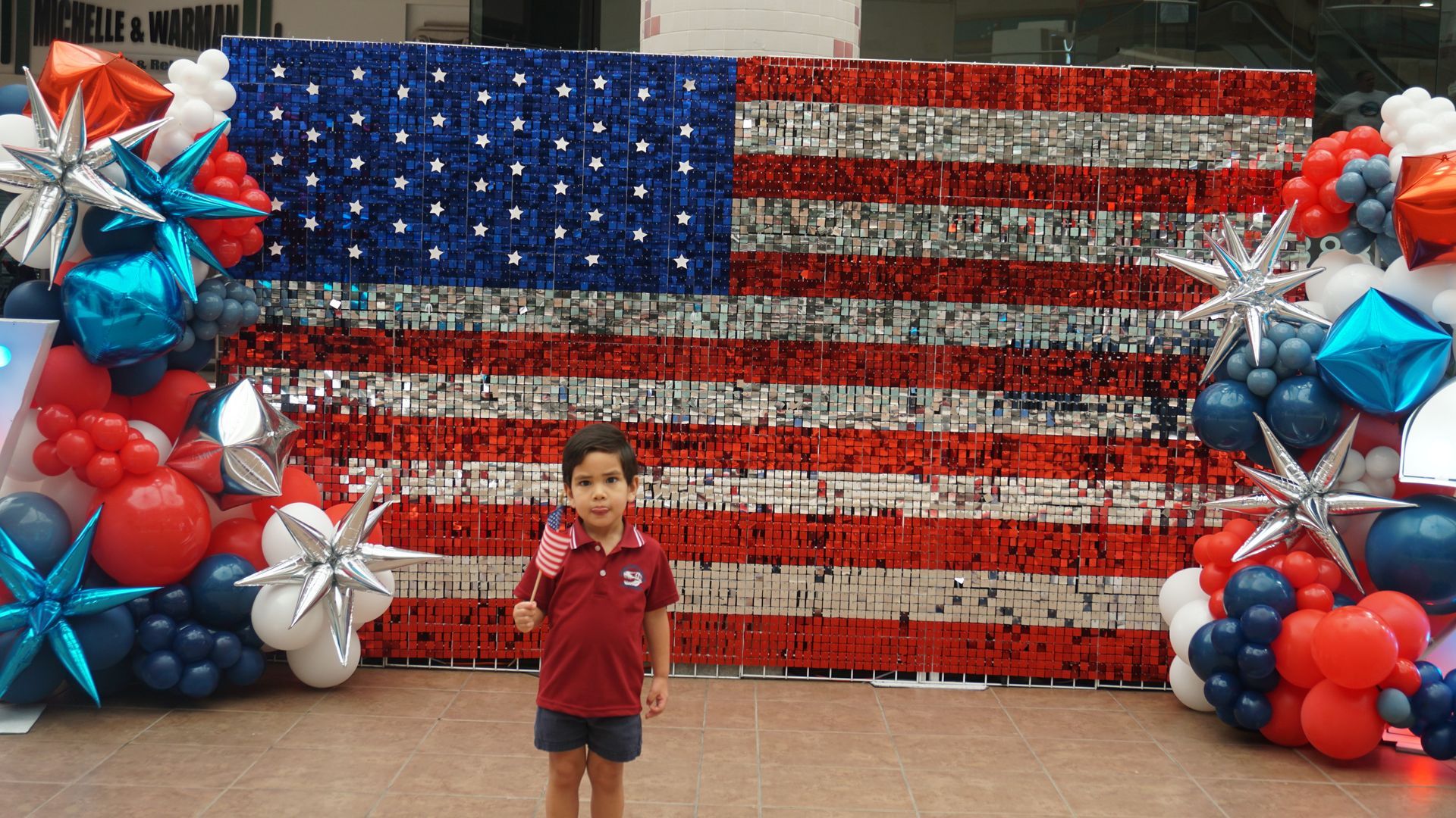 Child standing before a red, white, and blue balloon wall with a large American flag backdrop