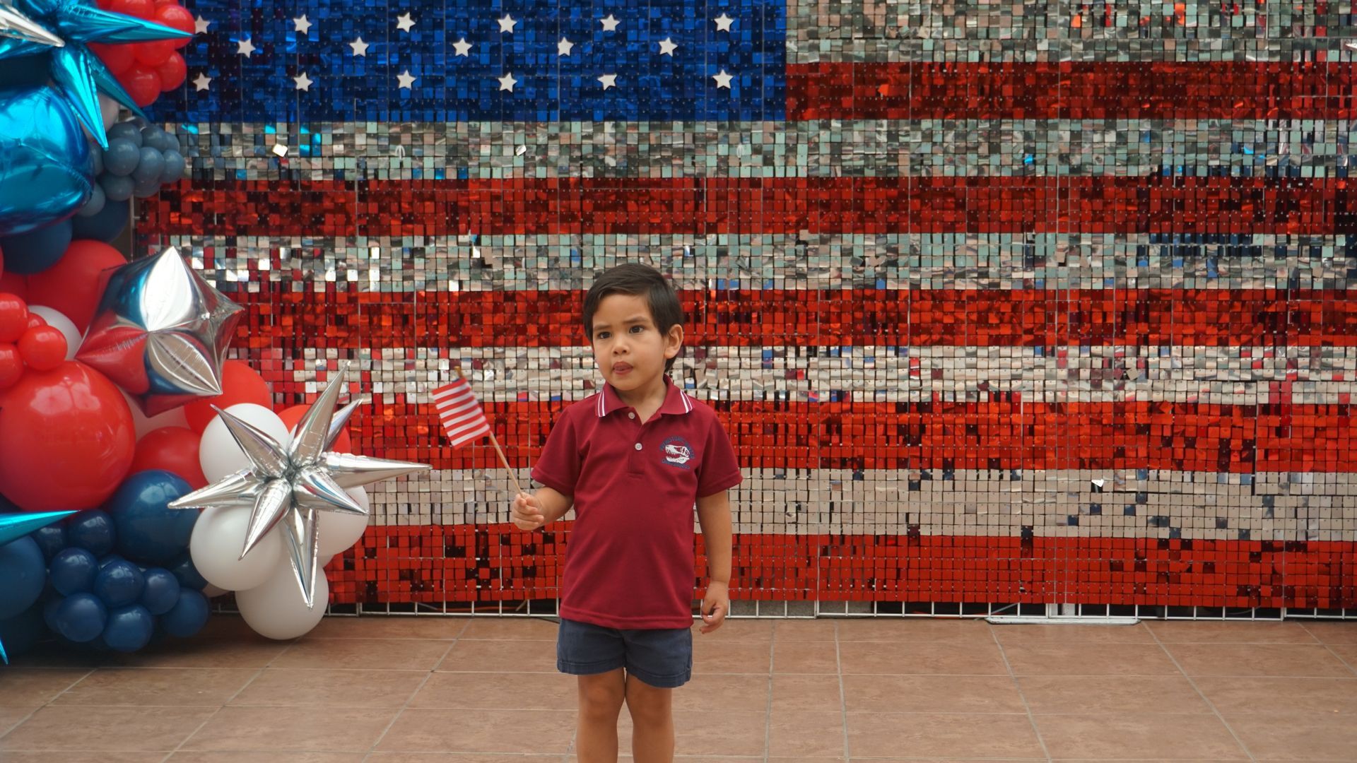 Child standing before a large American flag mural with red, white, and blue balloon decorations