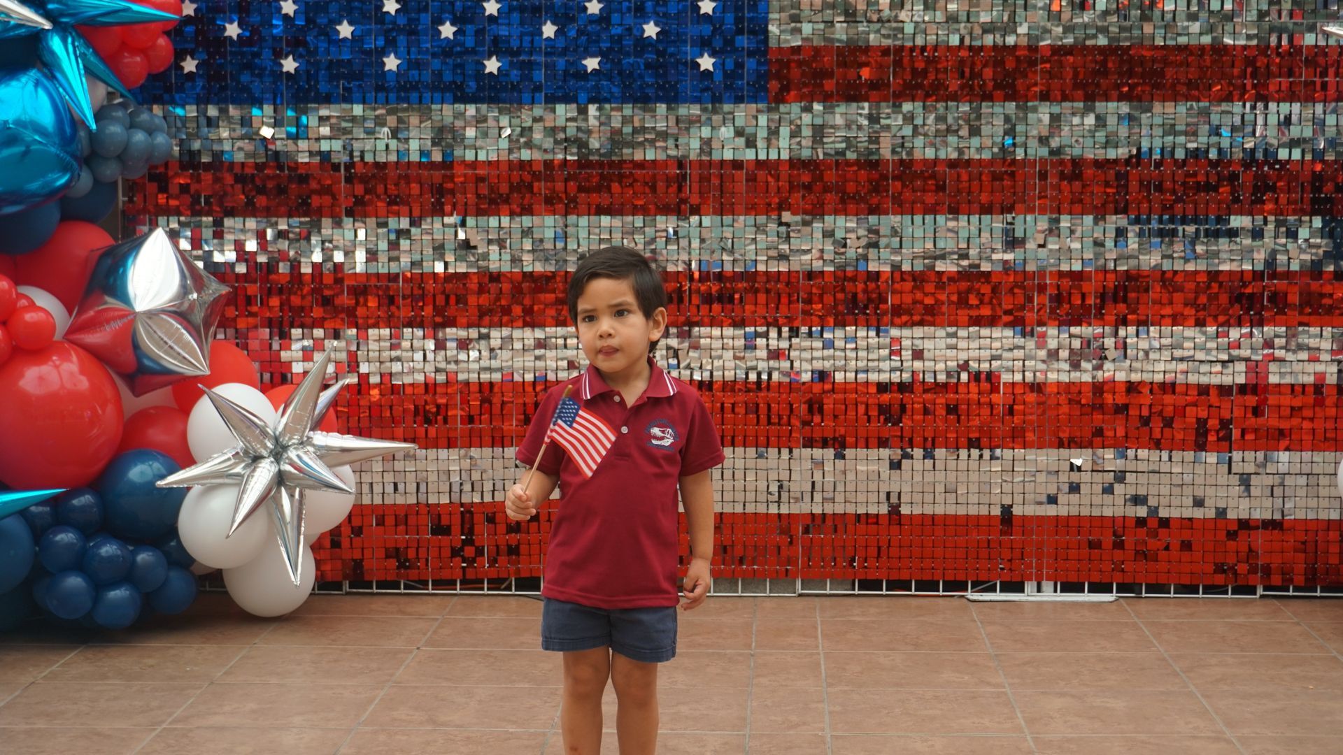 Boy holding a drink stands before a large American flag balloon display with red, white, and blue balloons