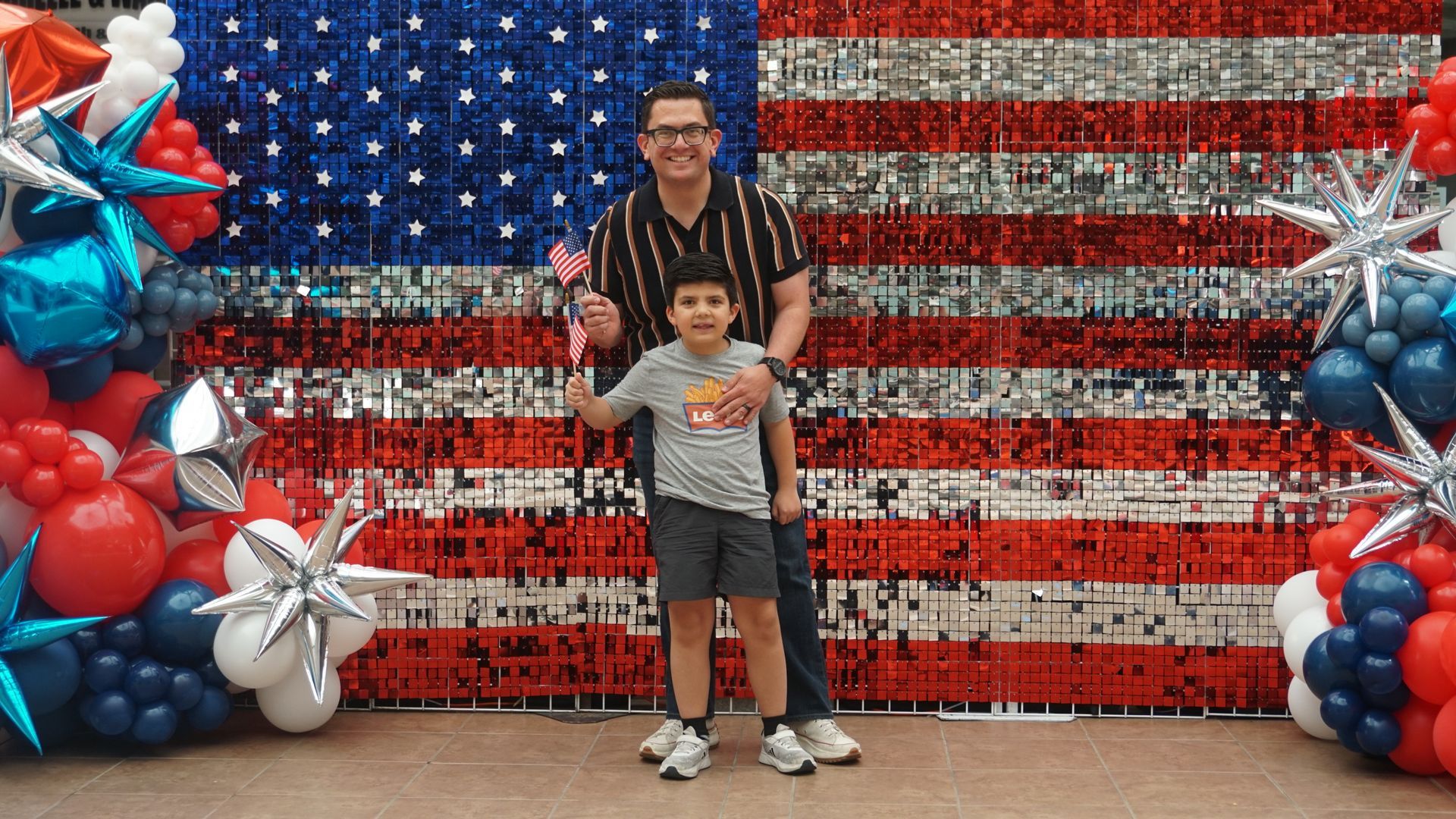 Two people standing before a red, white, and blue balloon display in front of an American flag backdrop
