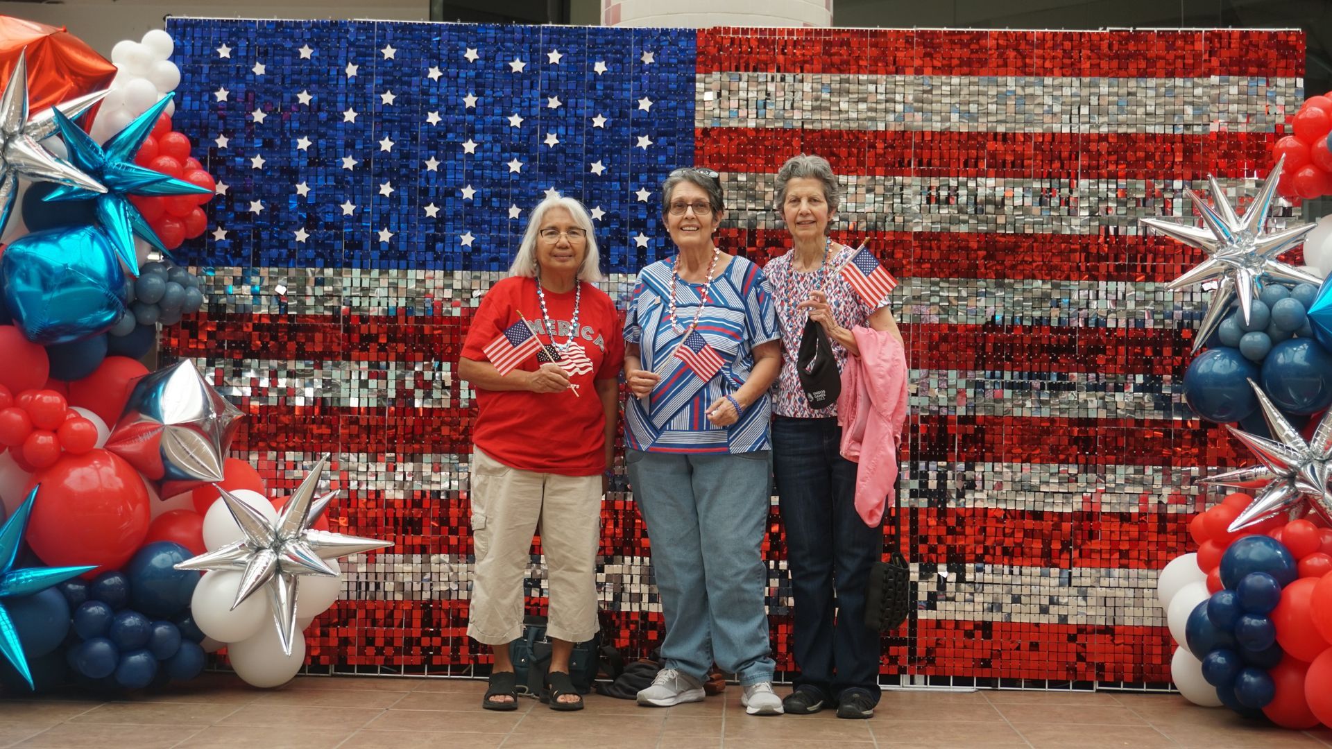 Three people posing before a giant American flag backdrop with red, white, and blue balloon columns.