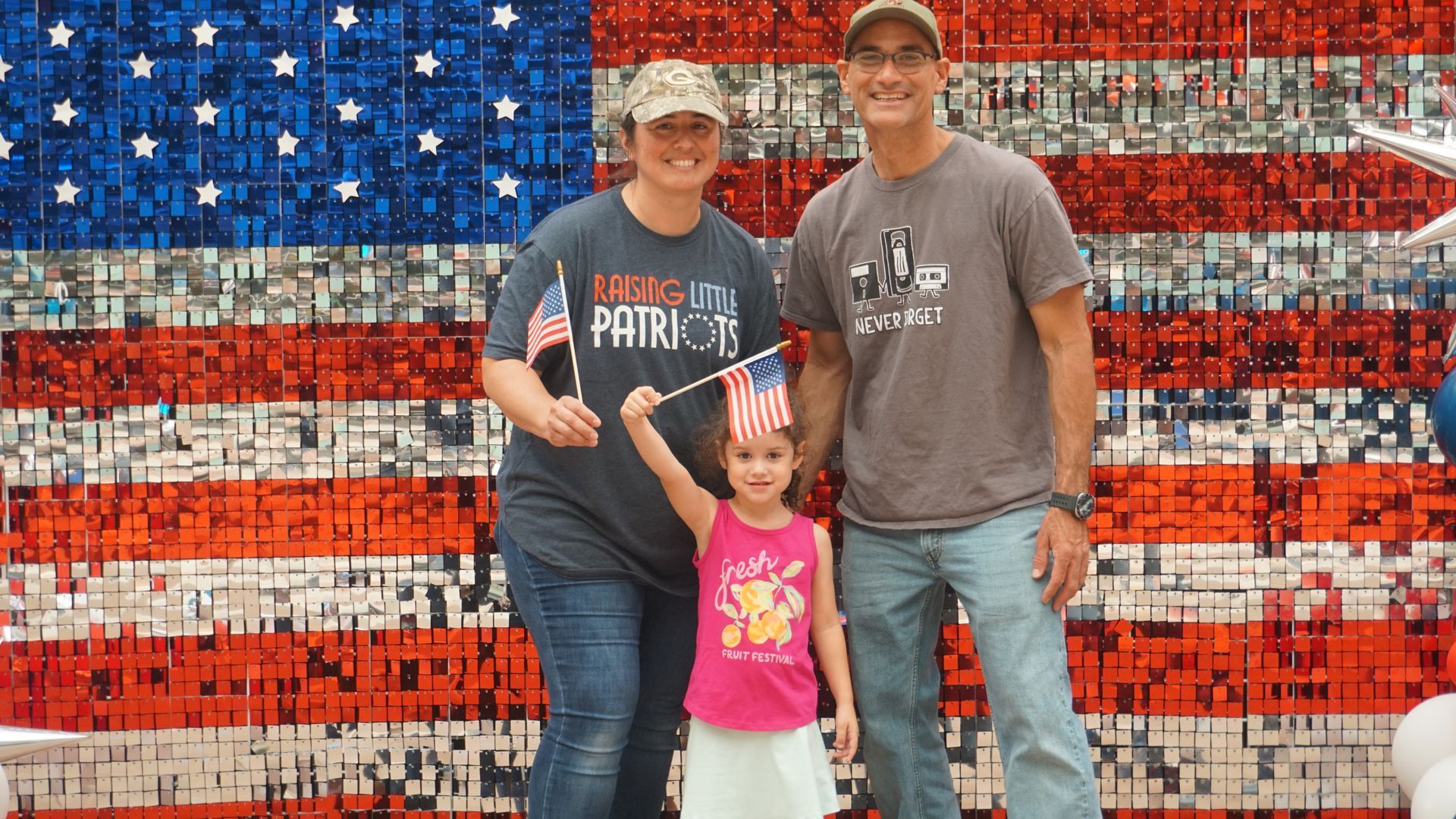 Three people posing in front of an American flag mural, with a child wearing a star headband in front.