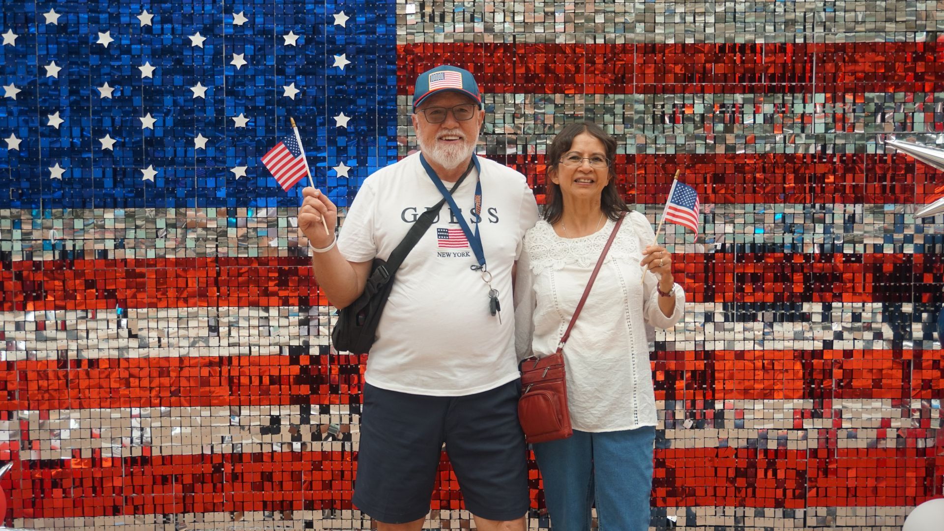 Two people posing in front of a large American flag made of red, white, and blue bottle caps