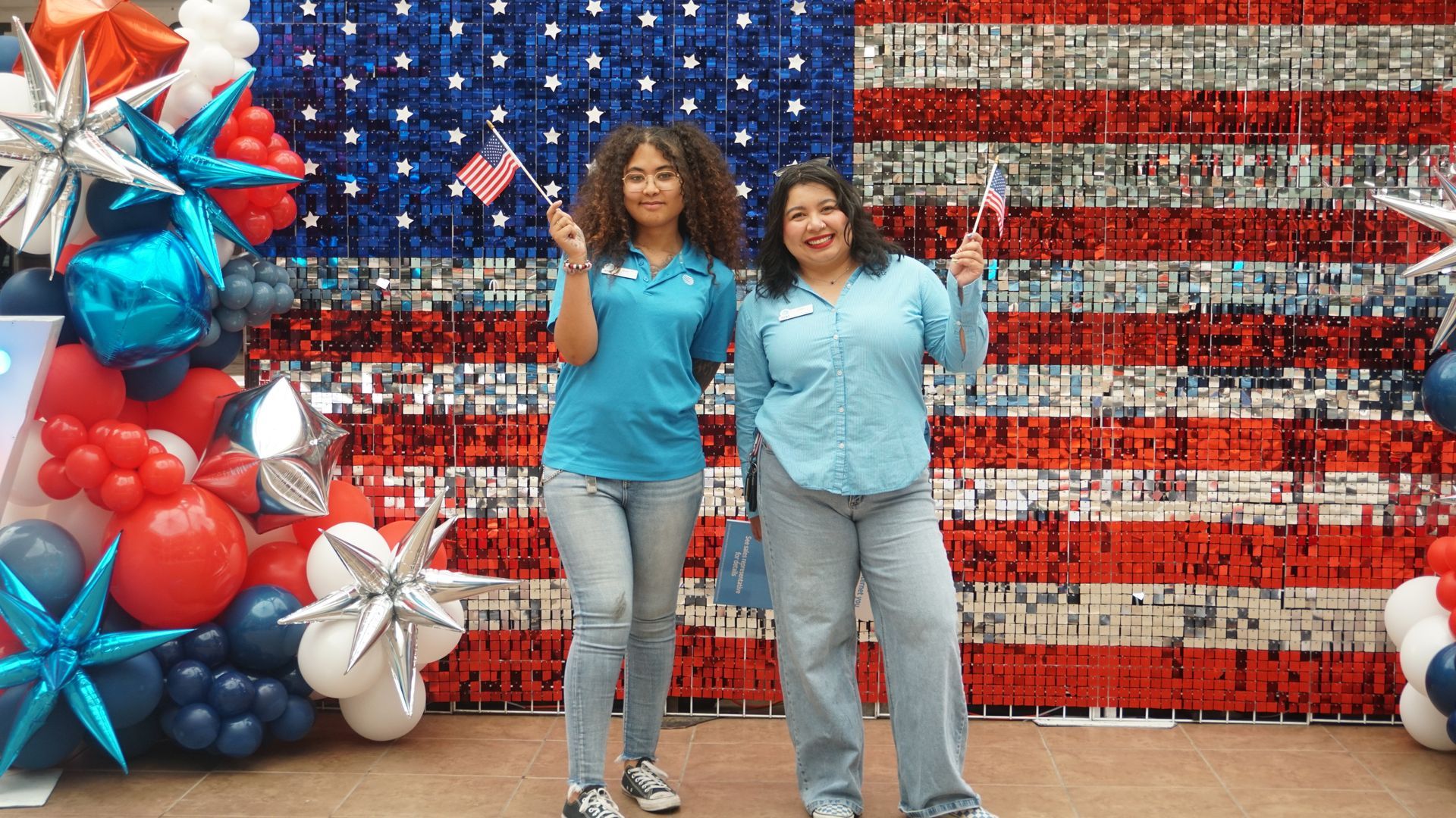 Two women standing before a red, white, and blue balloon American flag backdrop, smiling and waving.