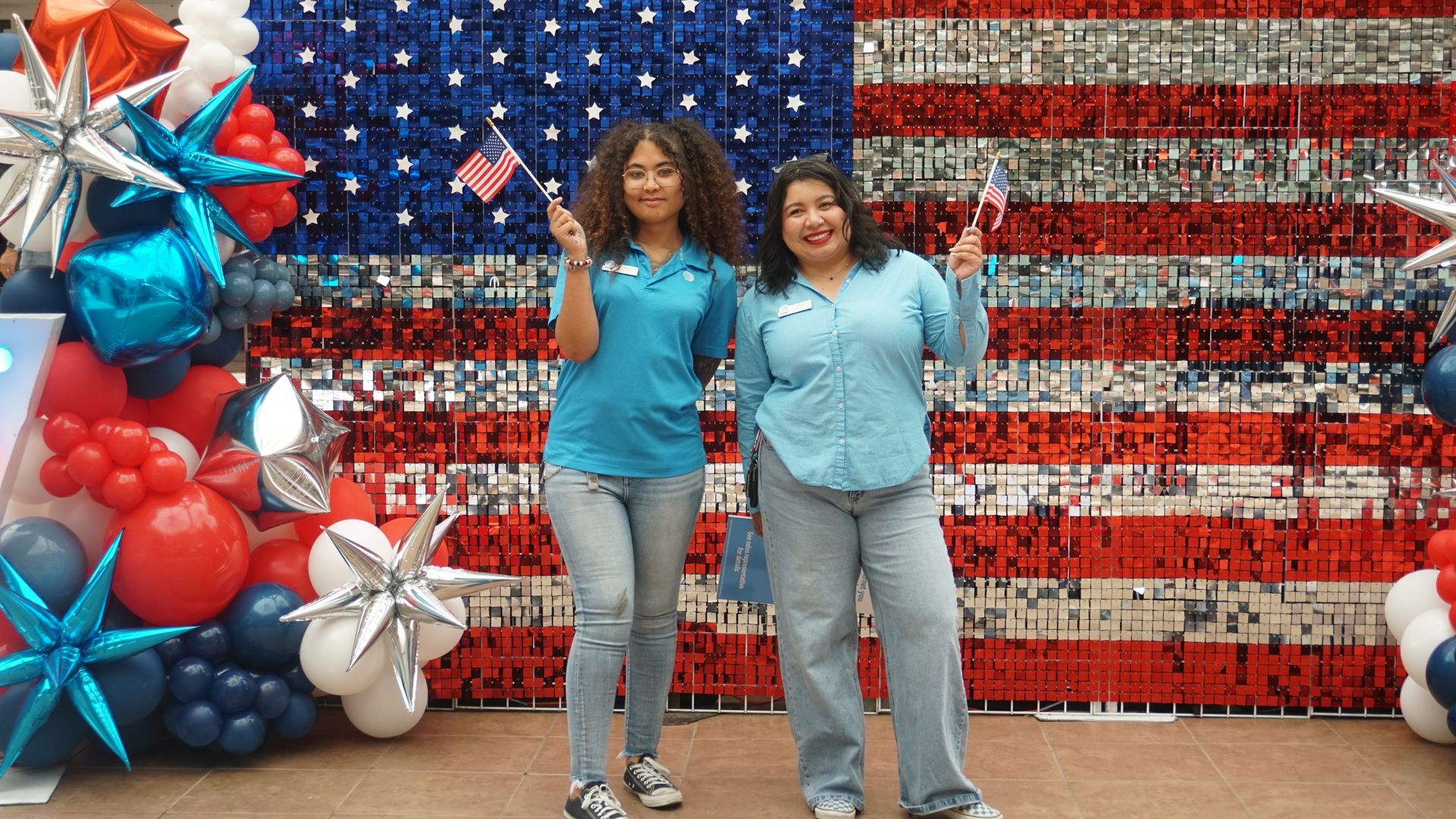 Two people in blue shirts stand before a red, white, and blue American flag display with balloons.