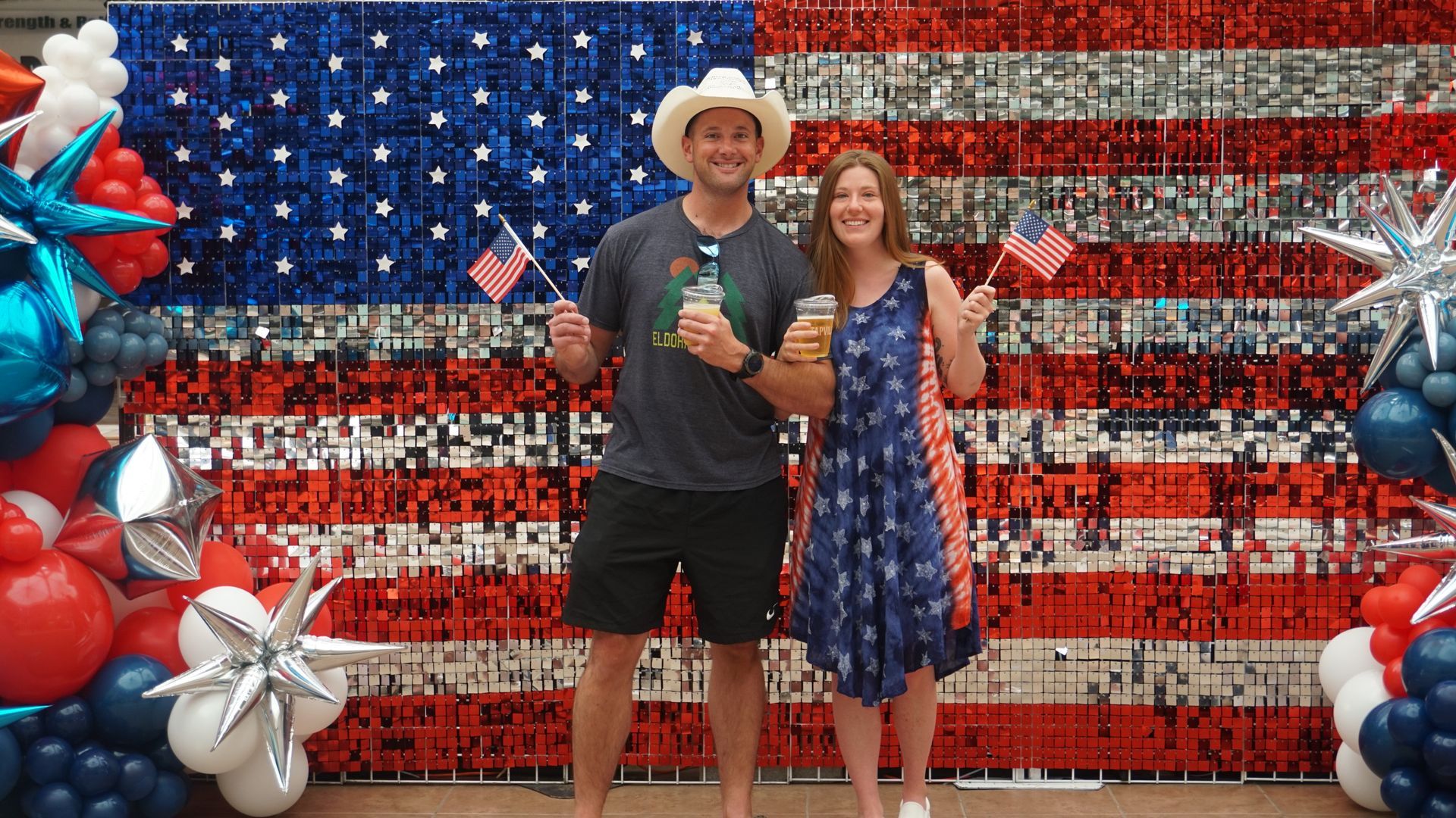 Two people posing in front of an American flag backdrop with red, white, and blue balloon decorations