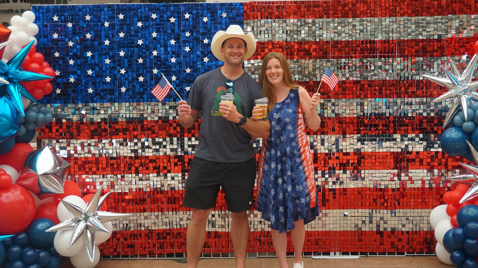 Two people posing in front of a large red, white, and blue American flag display with patriotic balloons