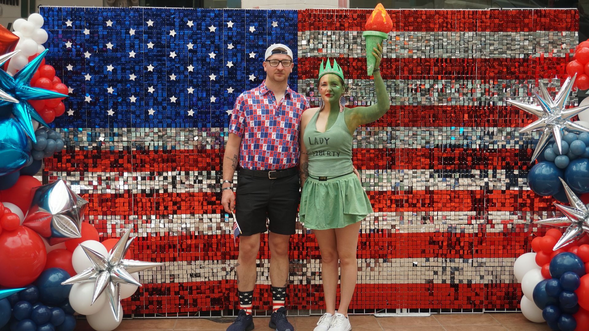Two people posing in front of an American flag display with red, white, and blue balloon decorations