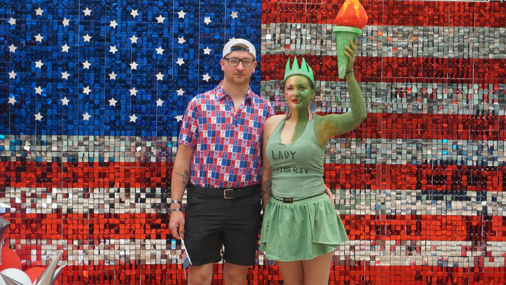 Two people pose before an American flag mural, one holding a lit torch aloft.