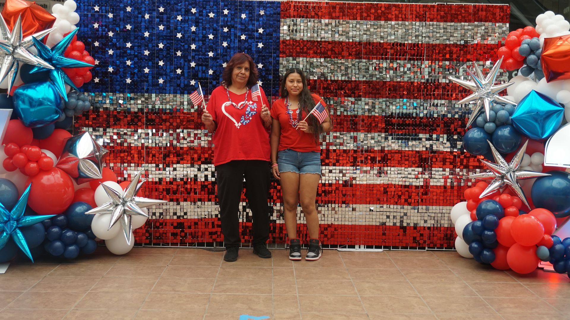 Two people posing before a patriotic red, white, and blue balloon-and-flag backdrop
