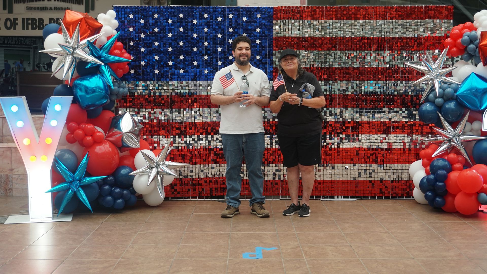 Two people stand in front of a patriotic red, white, and blue balloon backdrop with an American flag pattern.