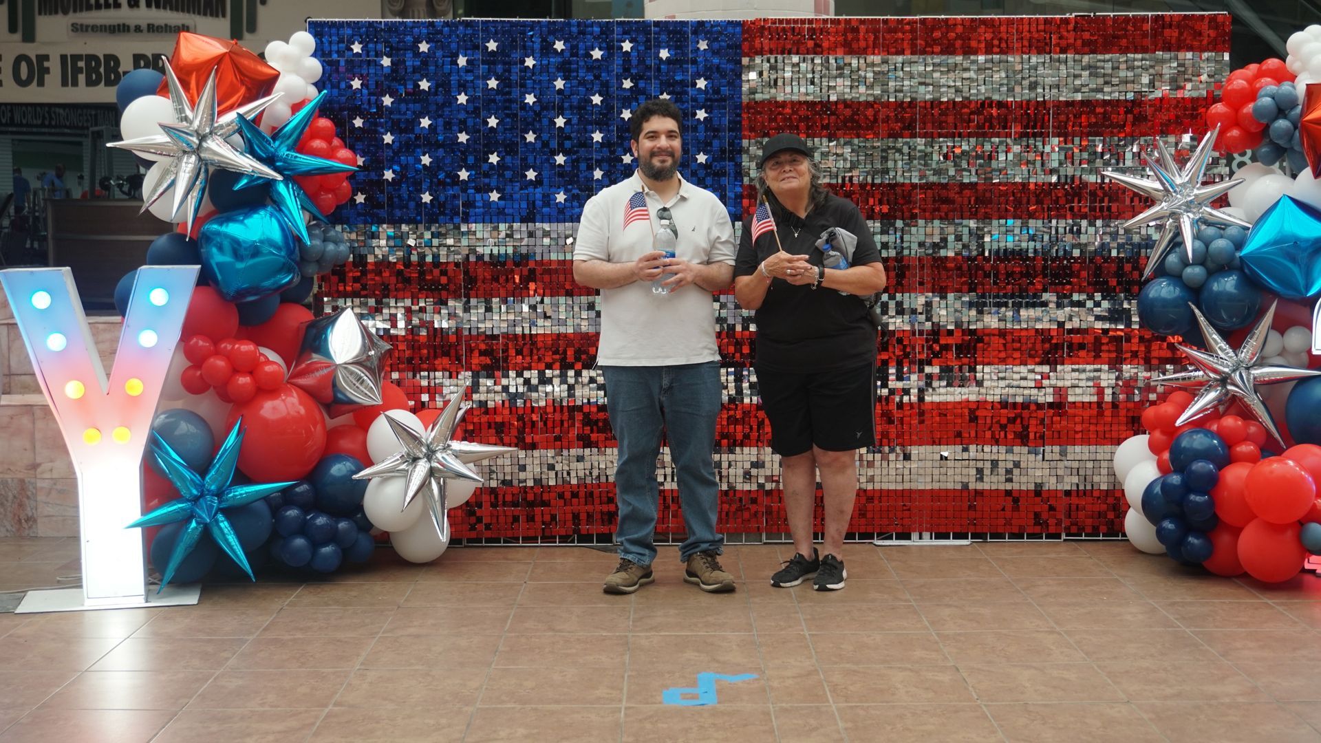 Two people stand before an American flag balloon backdrop, holding a sign at an event.