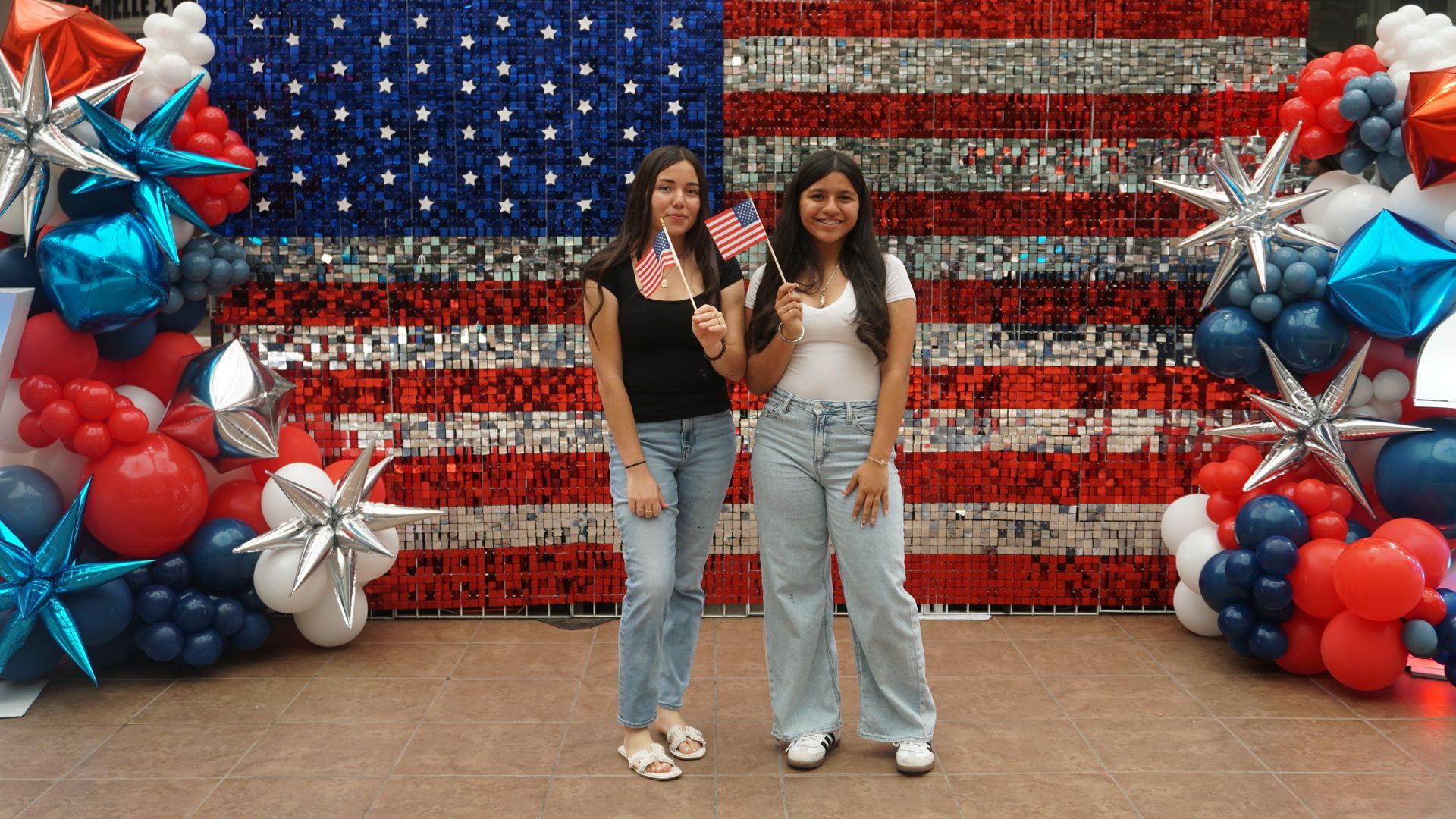 Two people posing before a red, white, and blue patriotic balloon backdrop