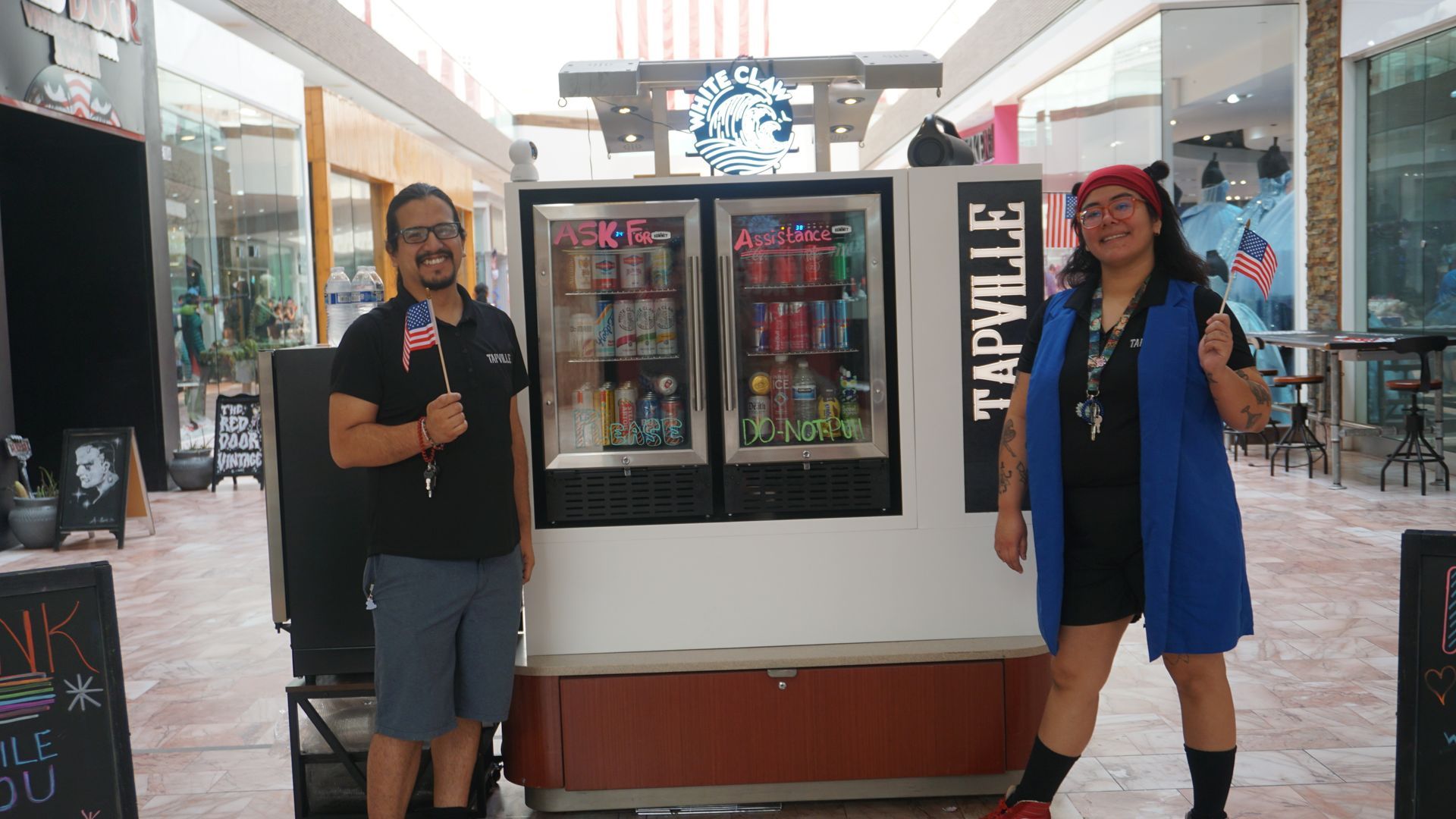Two people stand beside a small truck display in a mall, holding drinks and smiling.