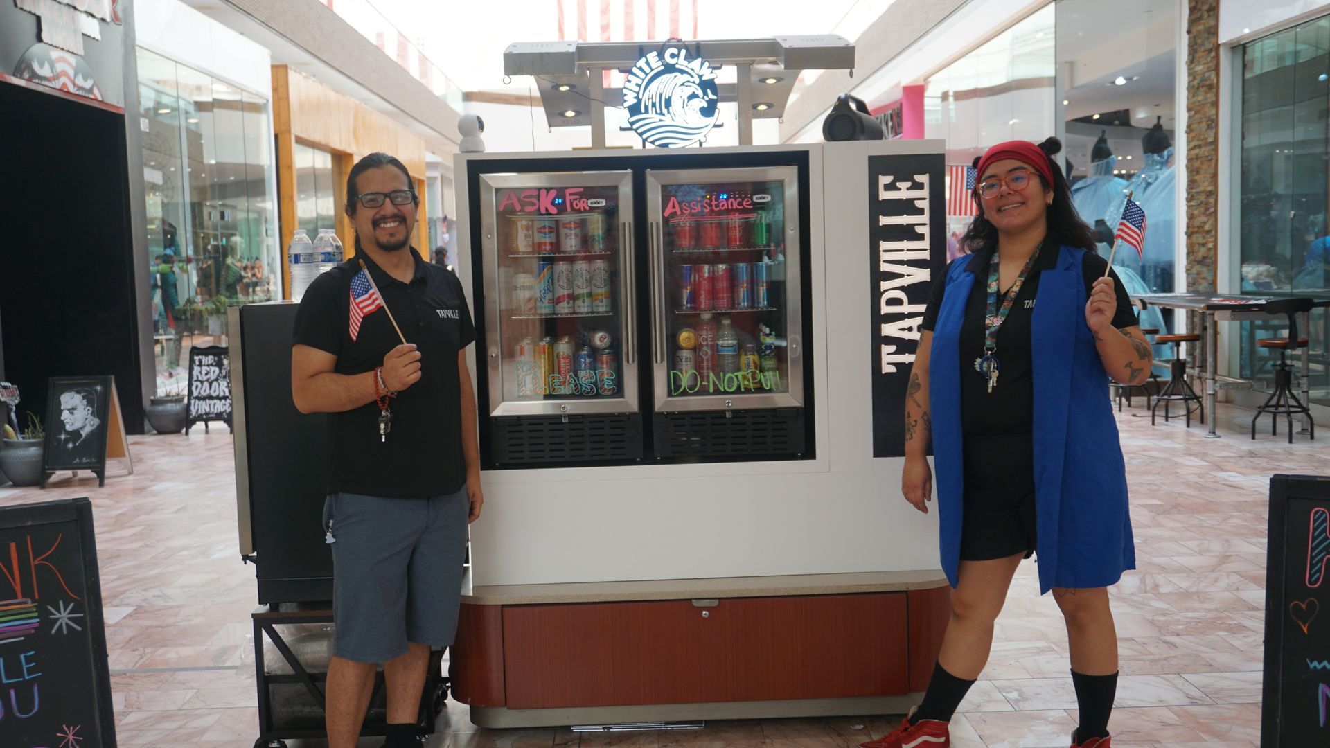 Two people stand beside a vending machine in a shopping mall corridor.