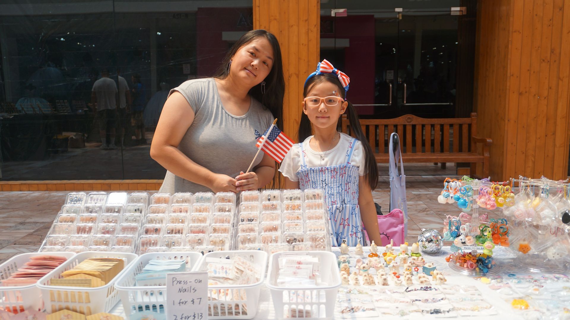 Two people behind a table of baked goods at an outdoor market, holding small American flags.