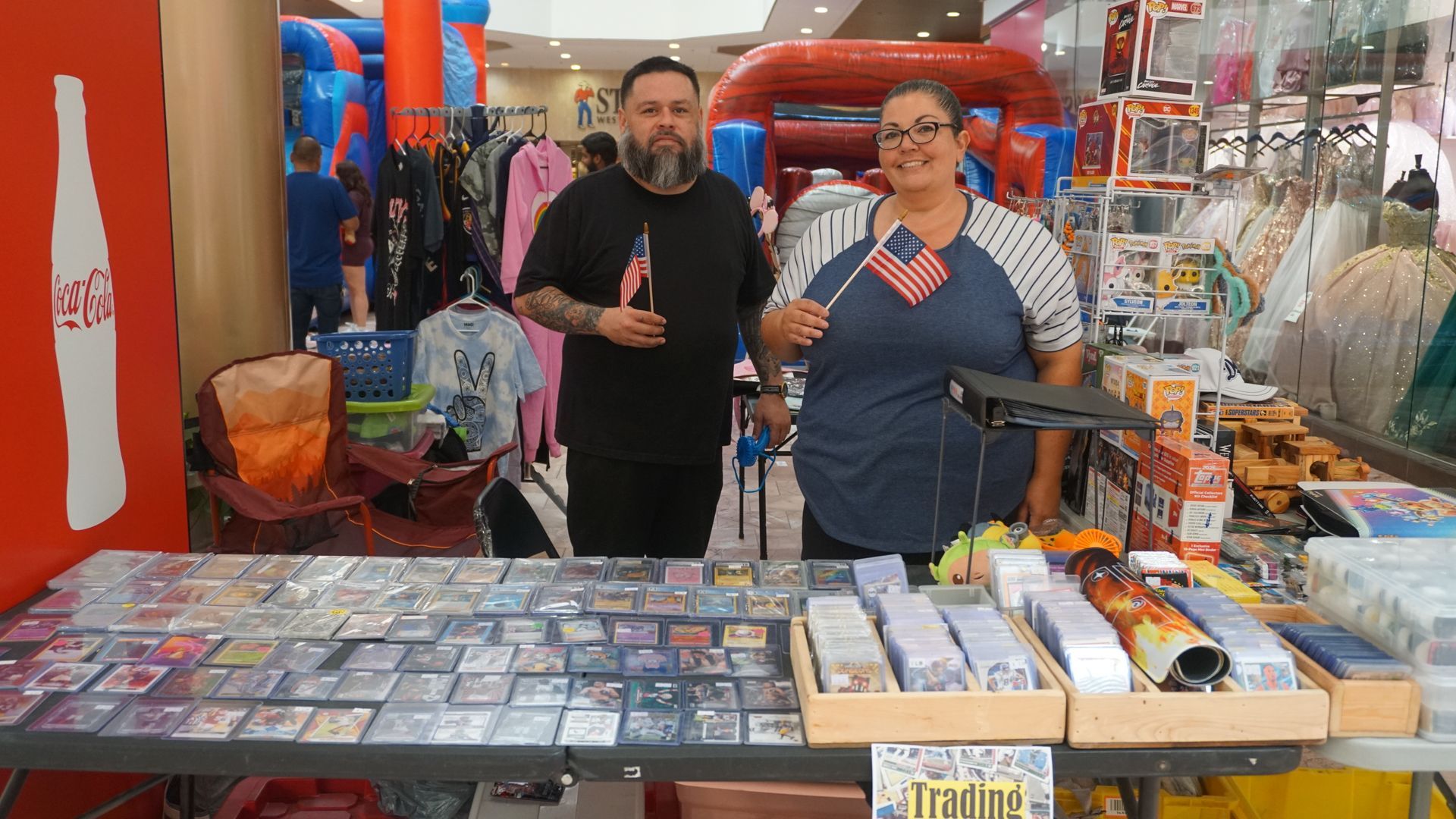 Two people behind a market stall with books and small items, one holding a microphone in a colorful indoor market