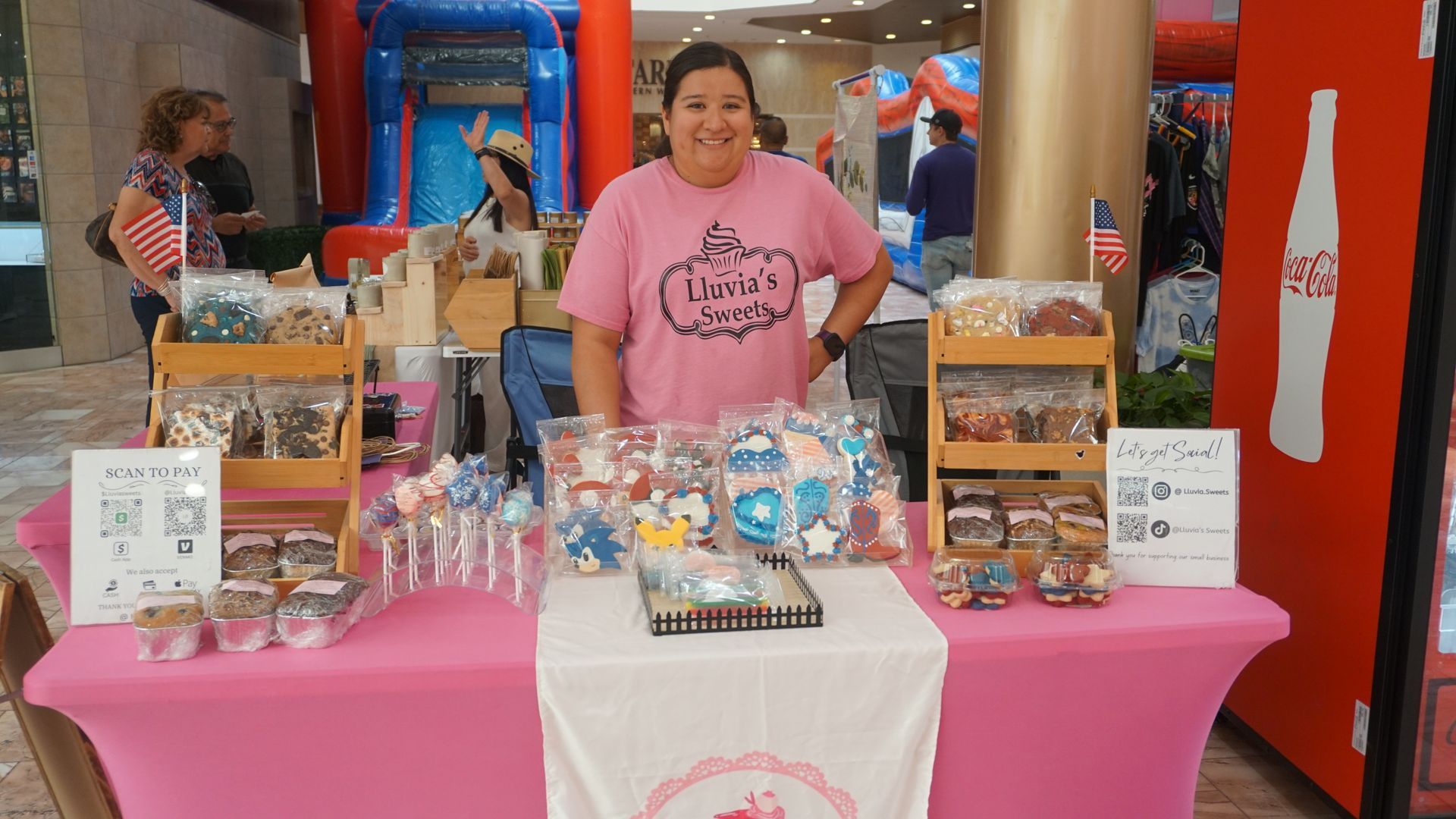 Woman behind pink market table displaying assorted treats and drinks indoors